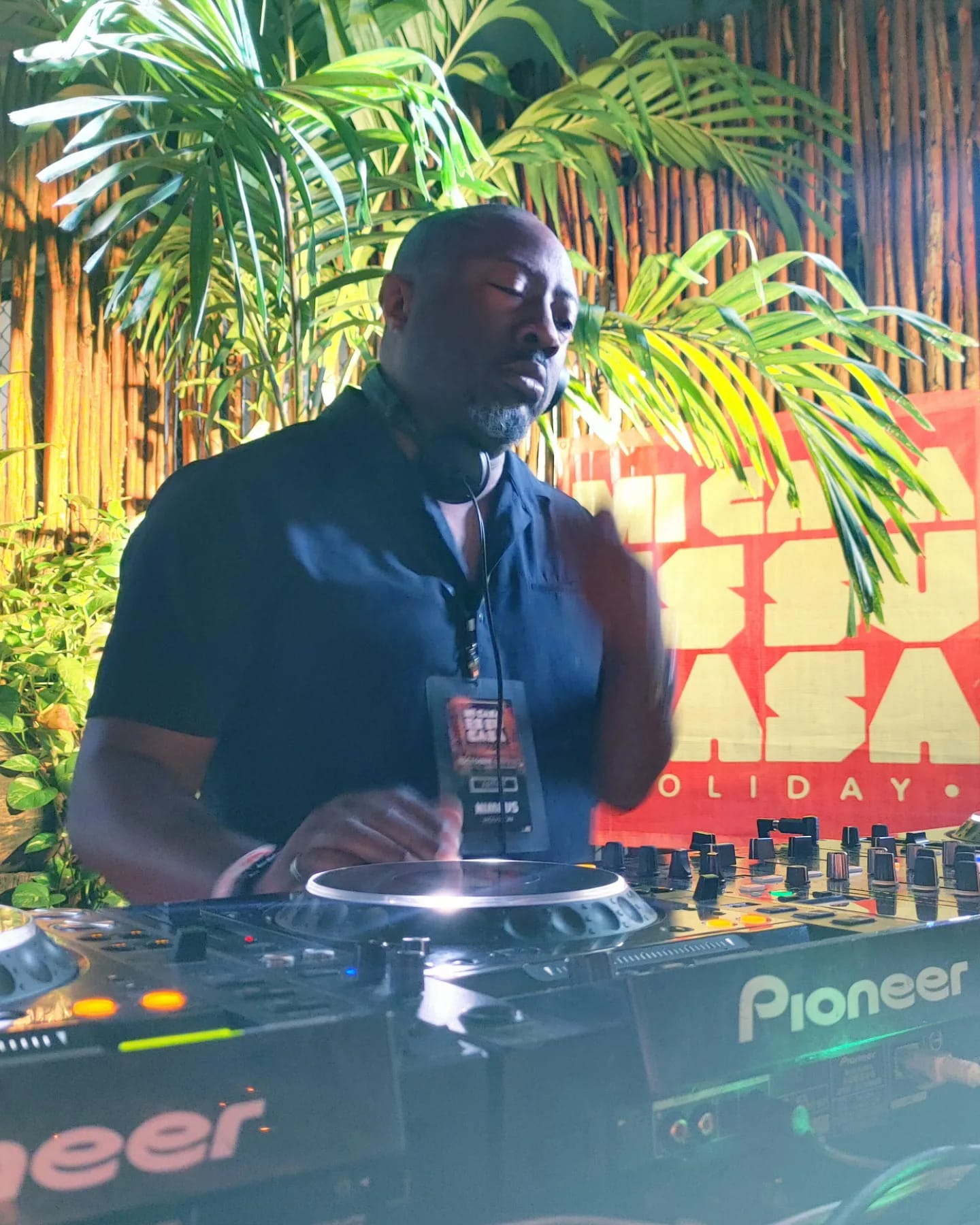 Person DJing outdoors with green plants and a red backdrop, wearing a dark shirt, holding a microphone, and operating DJ equipment.