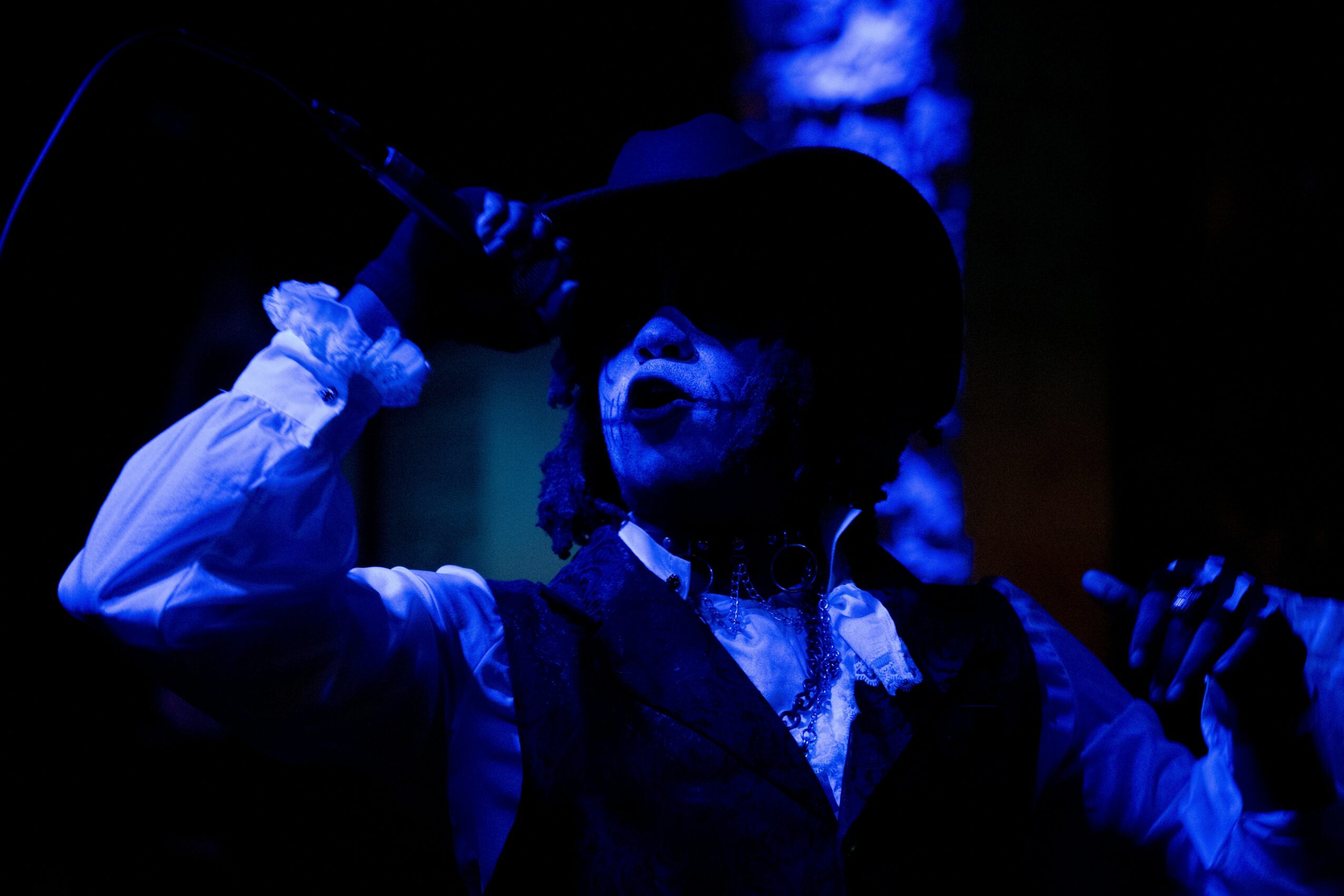 Person singing into a microphone, wearing a hat and vest, with a dark background.