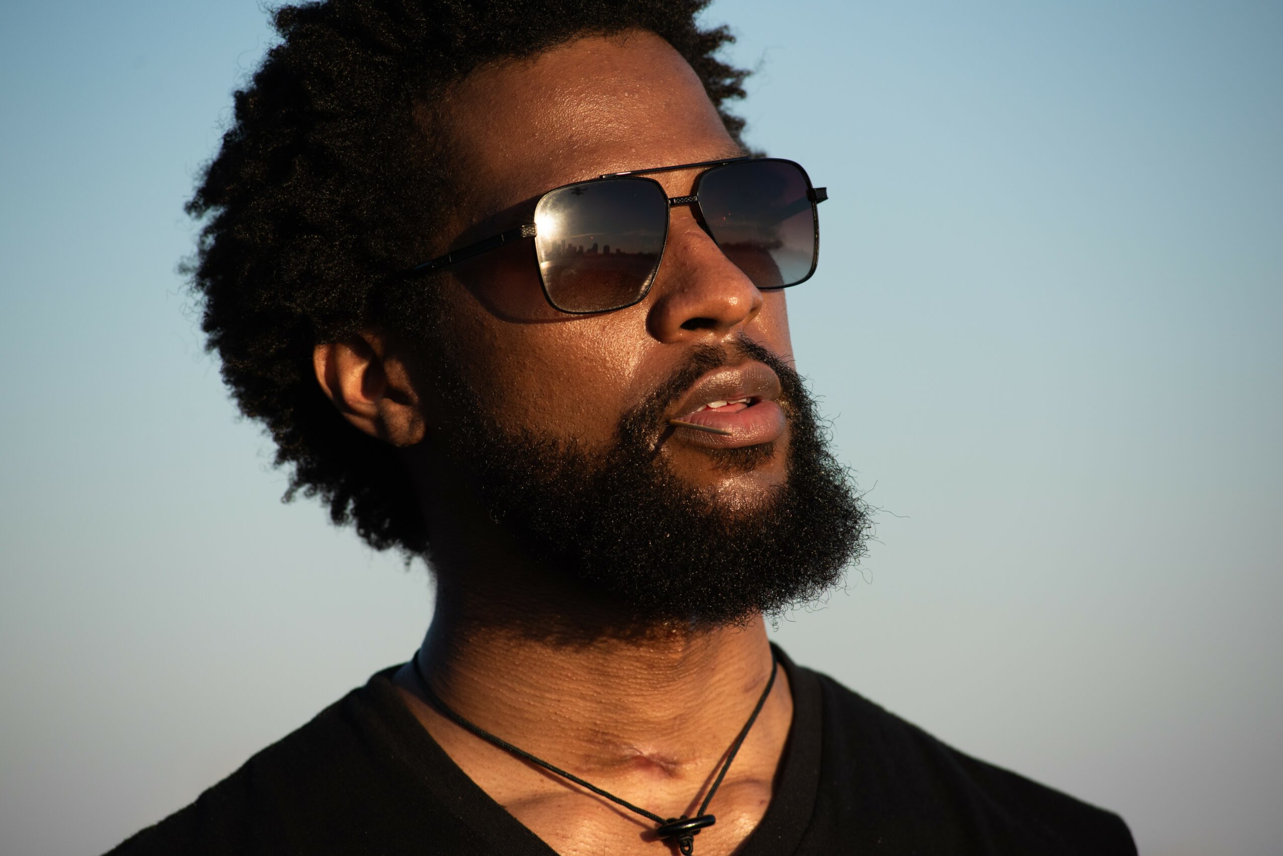 Man with sunglasses, beard, and curly hair outdoors against a clear sky, wearing a black shirt and necklaces.