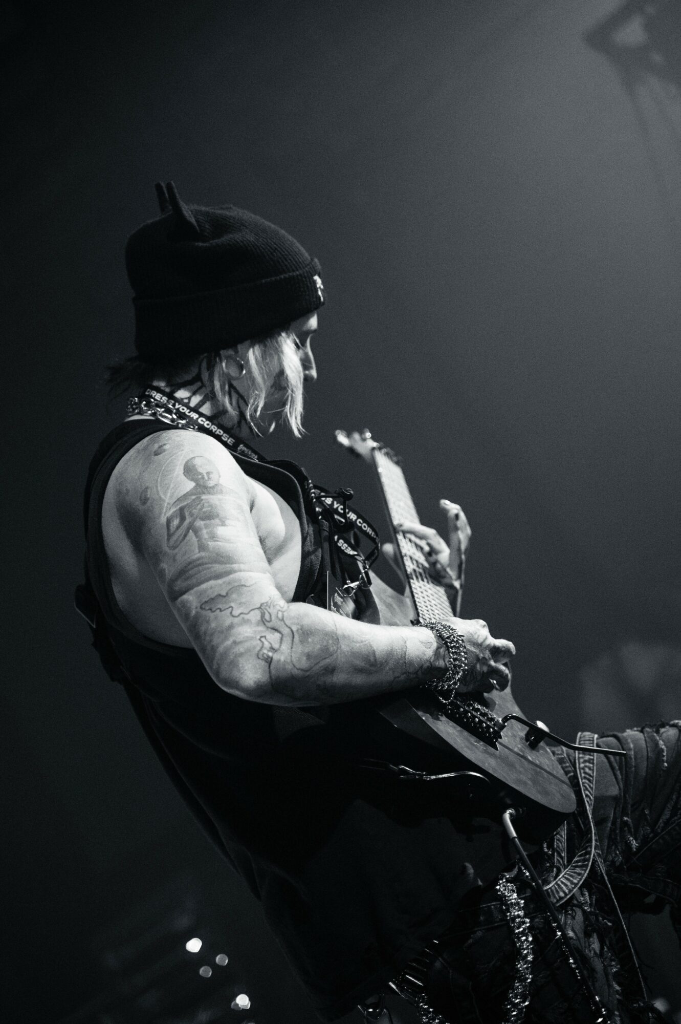 Musician playing guitar on stage, wearing a beanie and sleeveless shirt, with tattoos visible on arms, in black and white.