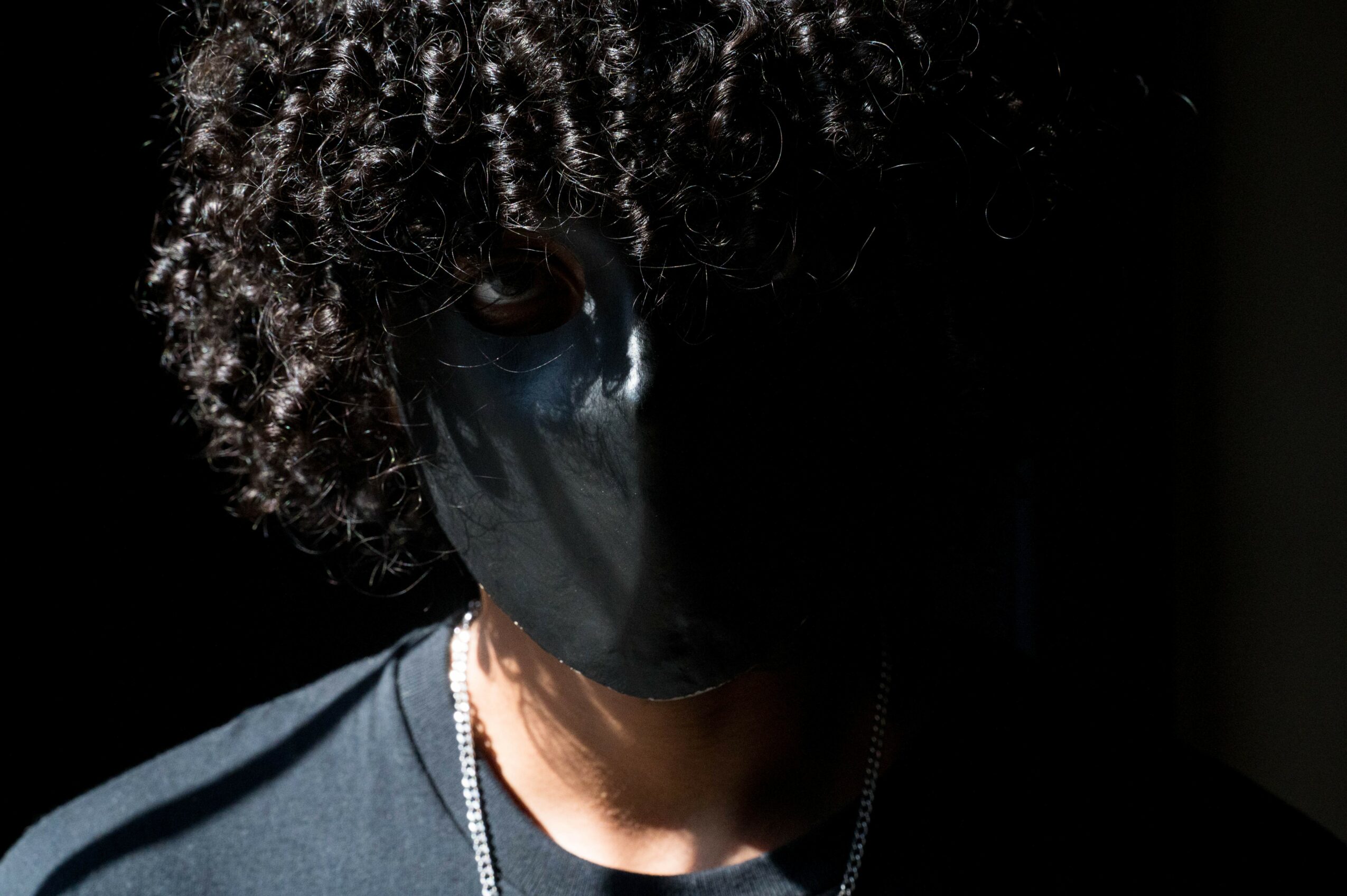 Person with curly hair wearing a black face mask and dark clothing, partially in shadow.