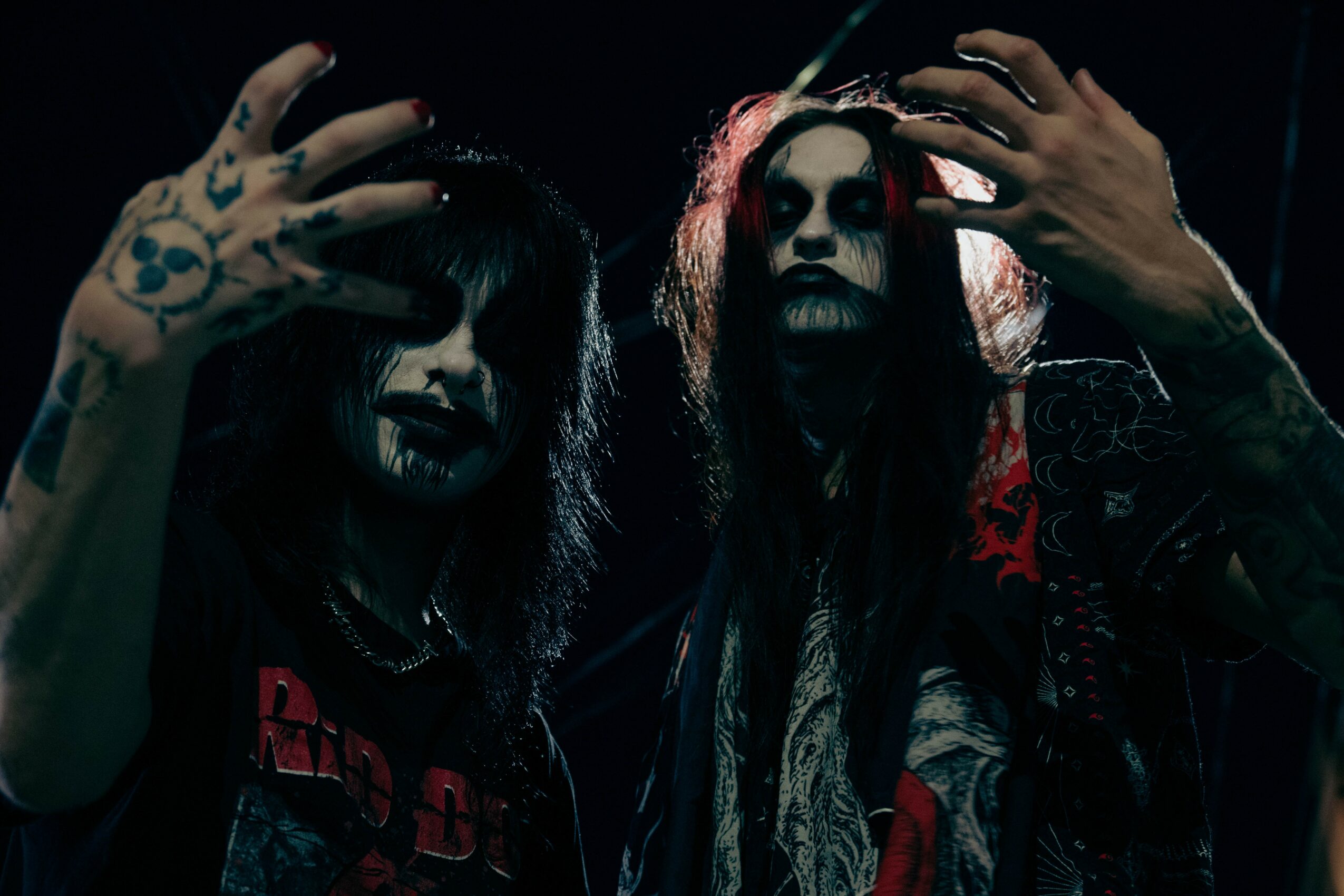Two people with dark makeup and long hair pose with hands raised, in a dark setting with dramatic lighting.