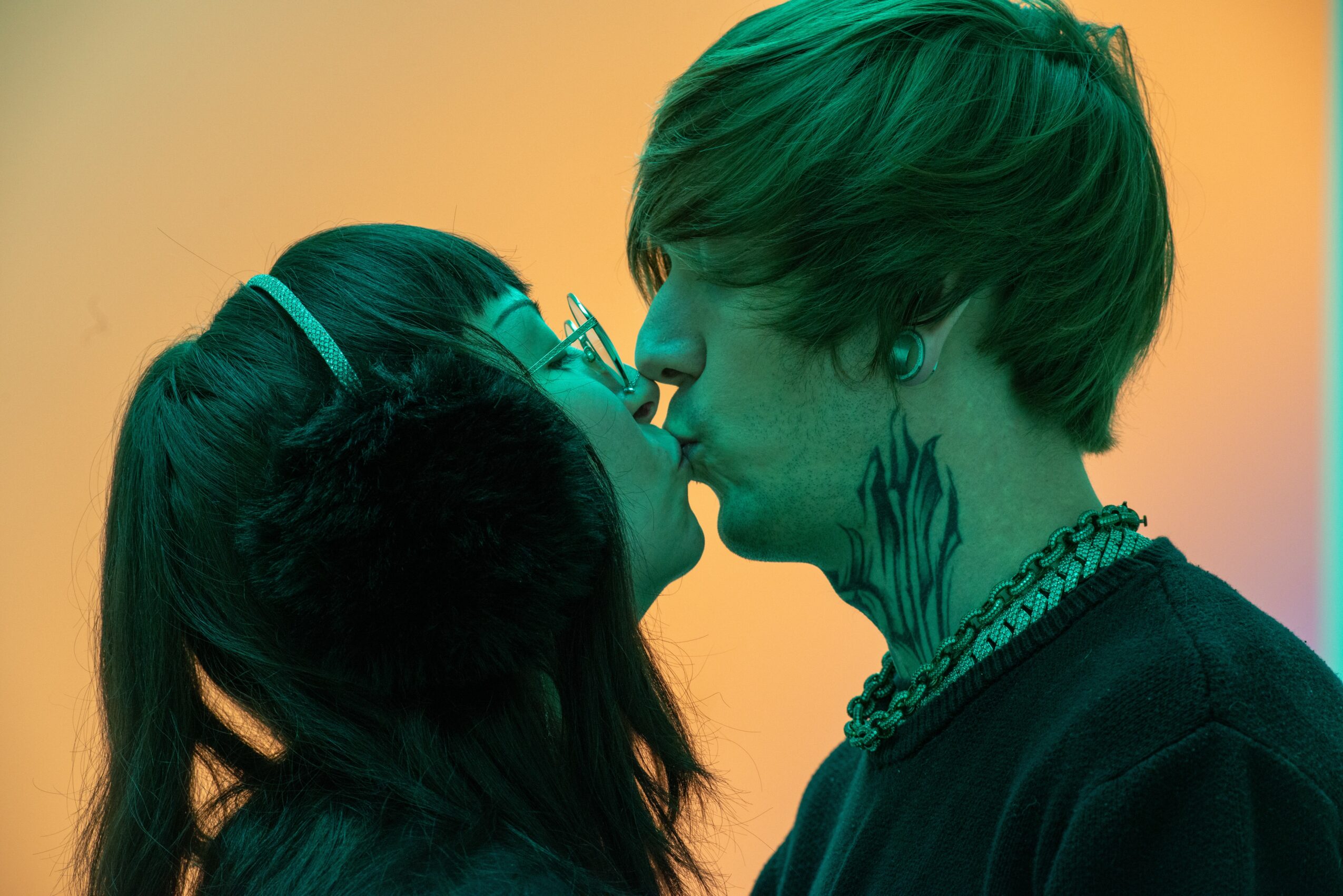 Two people with short hair and a ponytail are close, about to kiss, against a colorful background.