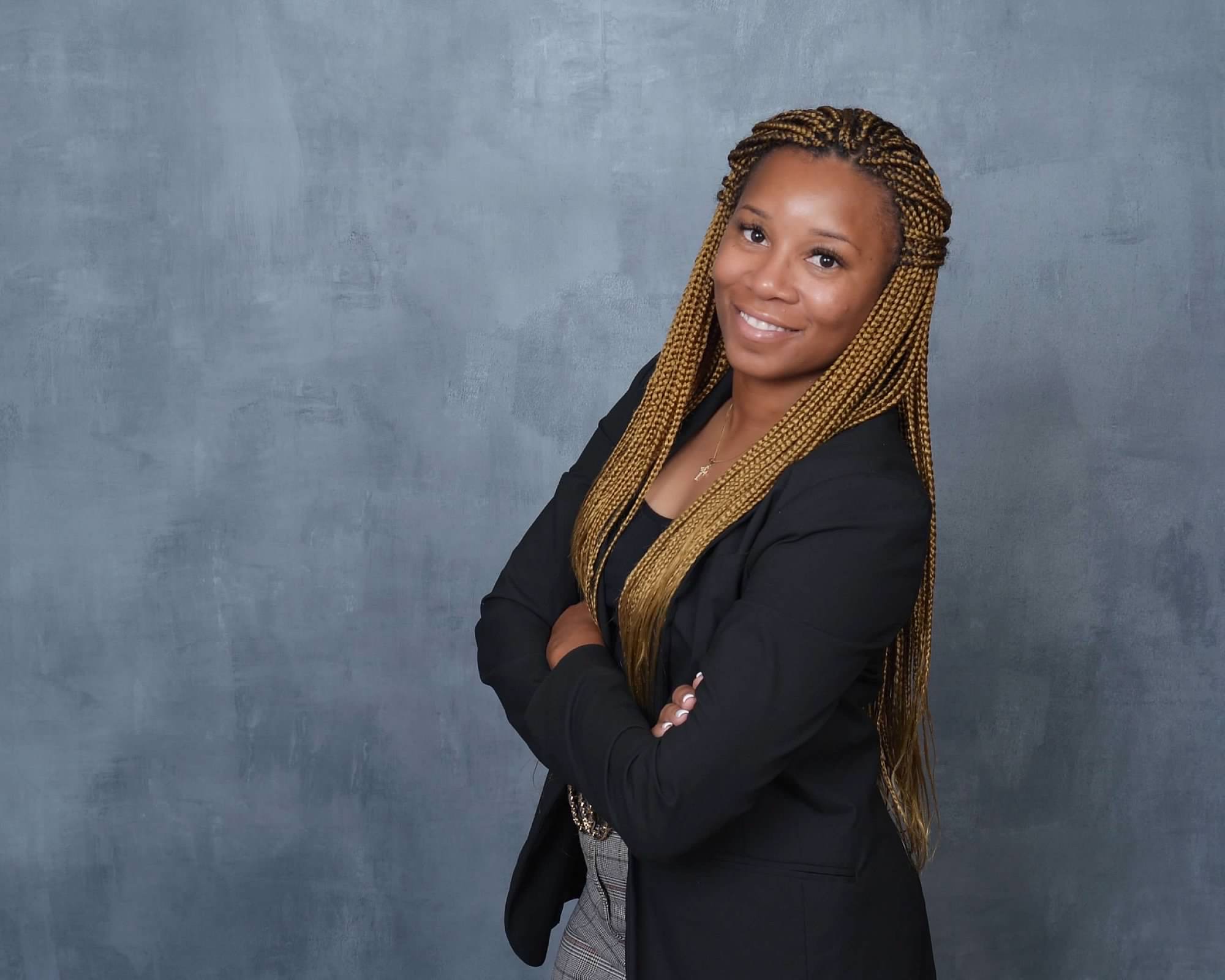 Woman with long braided hair smiling, arms crossed, wearing a black blazer against a gray background.