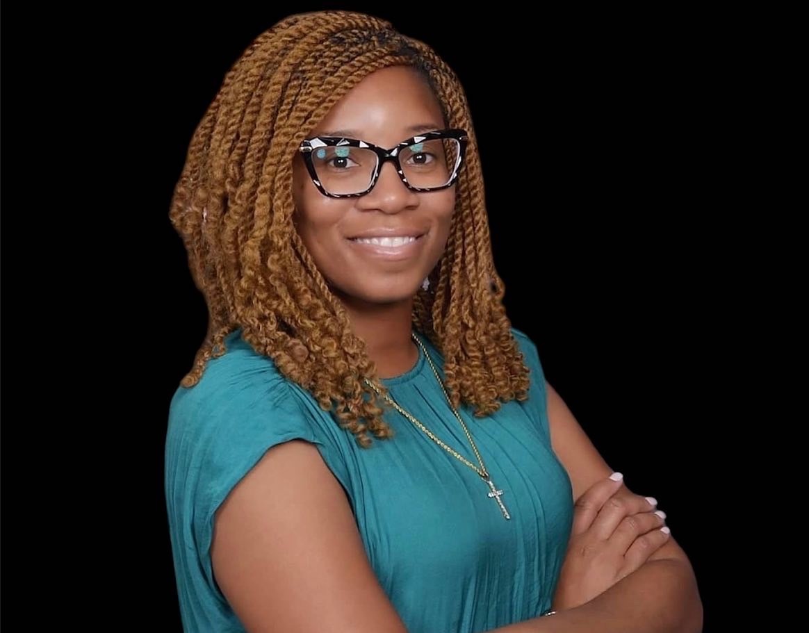 Woman with glasses and braided hair smiling, wearing a blue top and cross necklace, arms crossed, black background.