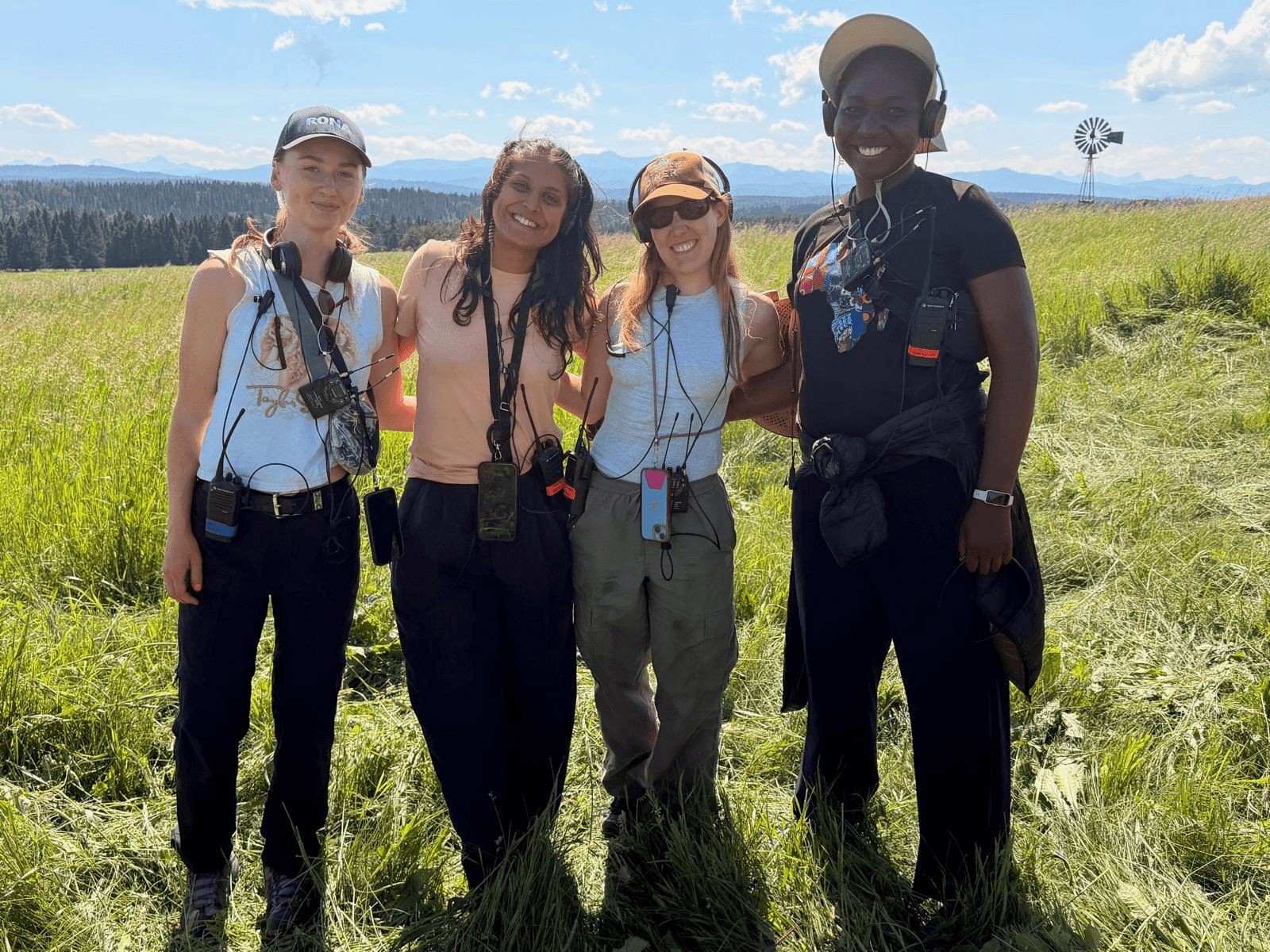Four people standing outdoors in a grassy field with mountains and blue sky in the background.
