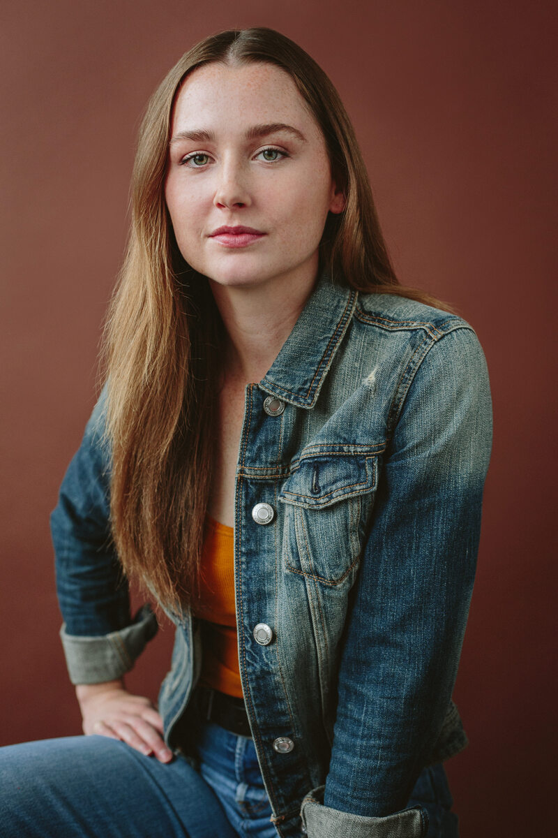 Young woman with long hair wearing a denim jacket, sitting against a brown background.