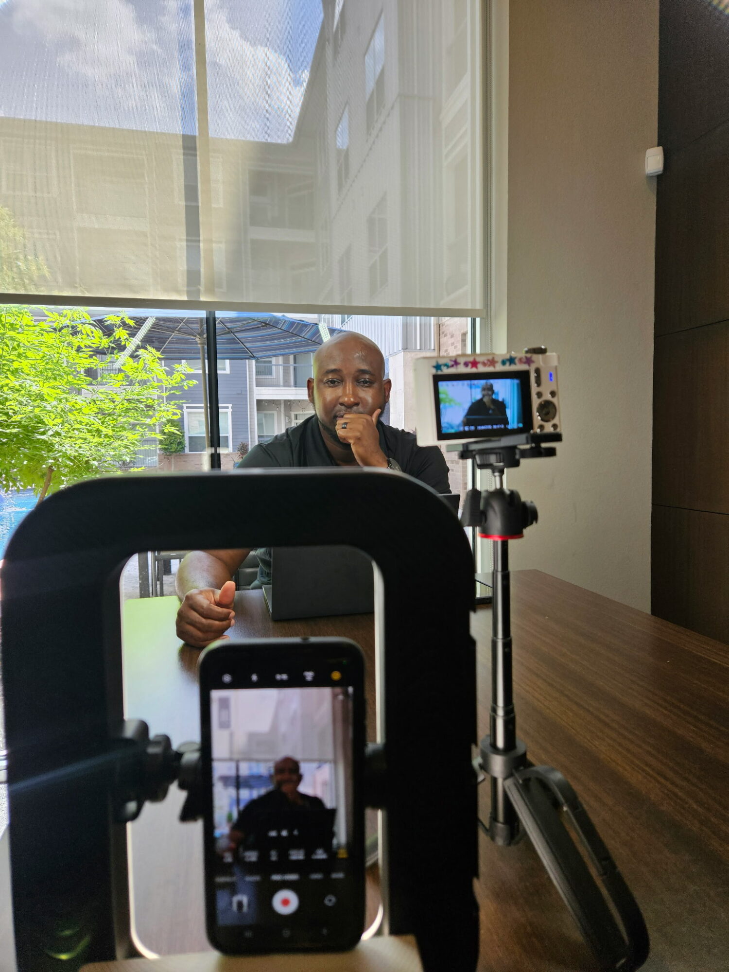 Person sitting at a table with camera and phone recording, outdoors with building and trees visible through window