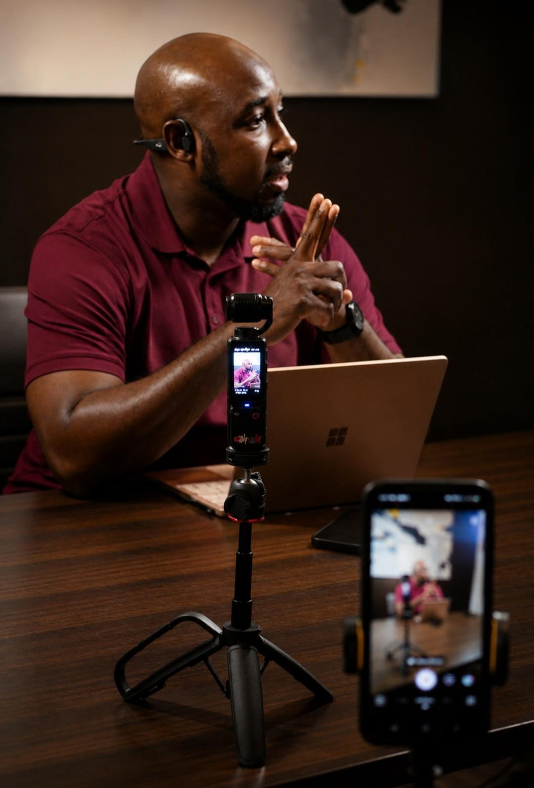 Man with bald head and beard, wearing a maroon shirt, sitting at a table with a laptop, holding hands together, and a smartphone on a tripod in front.