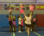 Group of cheerleaders performing a stunt with two lifted girls, one in pink dress, in an indoor gym with flags. Word count: 20