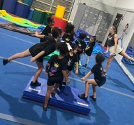 Group of children practicing gymnastics on a blue mat in a gym, with an instructor guiding them.