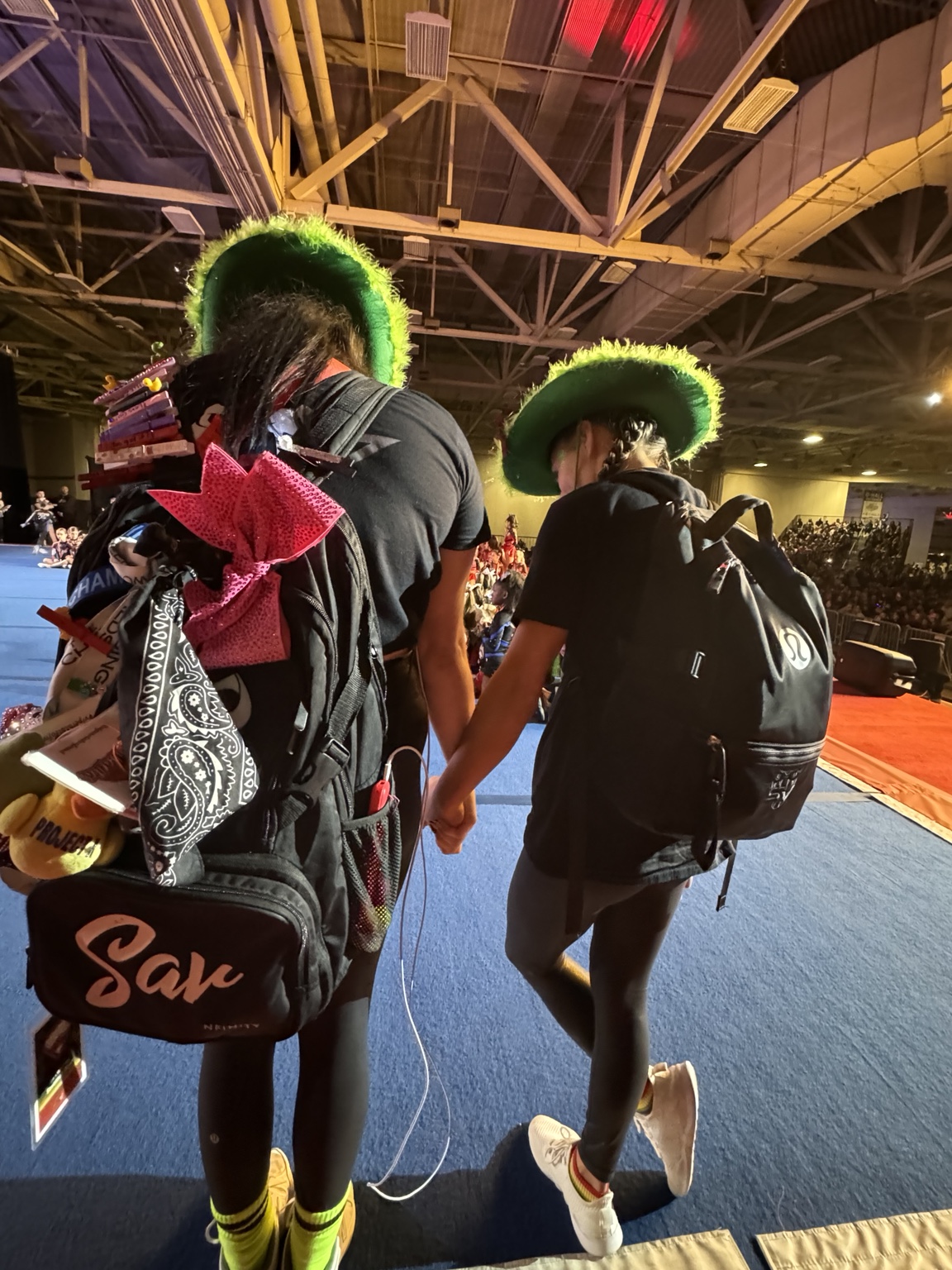 Two people wearing green hats and backpacks walking on a blue carpeted area indoors, with a large ceiling structure above.
