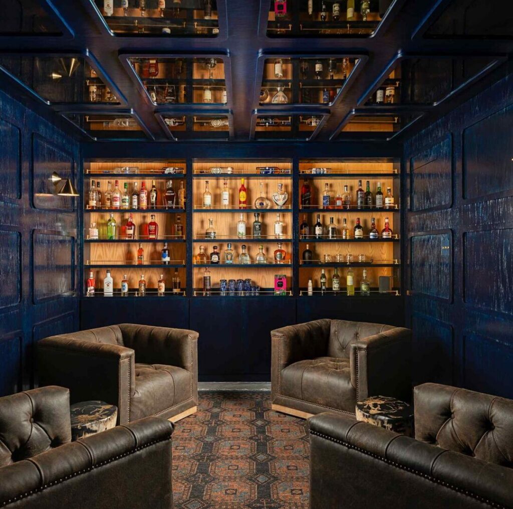 Bar with shelves of bottles, two armchairs, and a patterned carpet, viewed from the front, ceiling with glass panels.