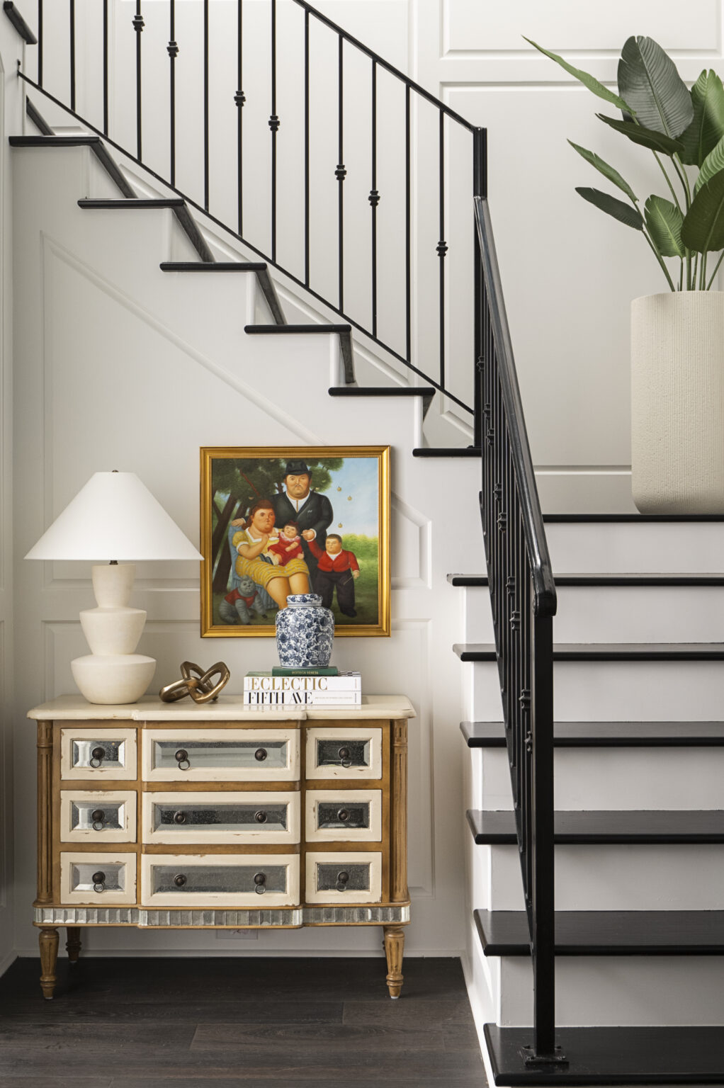 Interior staircase with black railing, a framed photo, a white lamp, and a large potted plant on a white wall, 20 words.