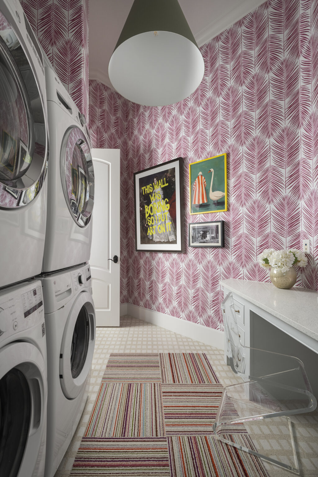 Laundry room with stacked washing machines, pink patterned wallpaper, framed artwork, a white cabinet, and a striped rug.