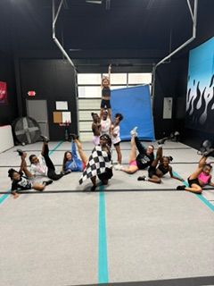 Group of people in a gym, some sitting and some standing, with a blue mat and black wall in the background.