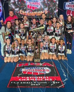 Group of young cheerleaders celebrating with a trophy, medals, and confetti on a stage, with a banner and championship sign behind them.