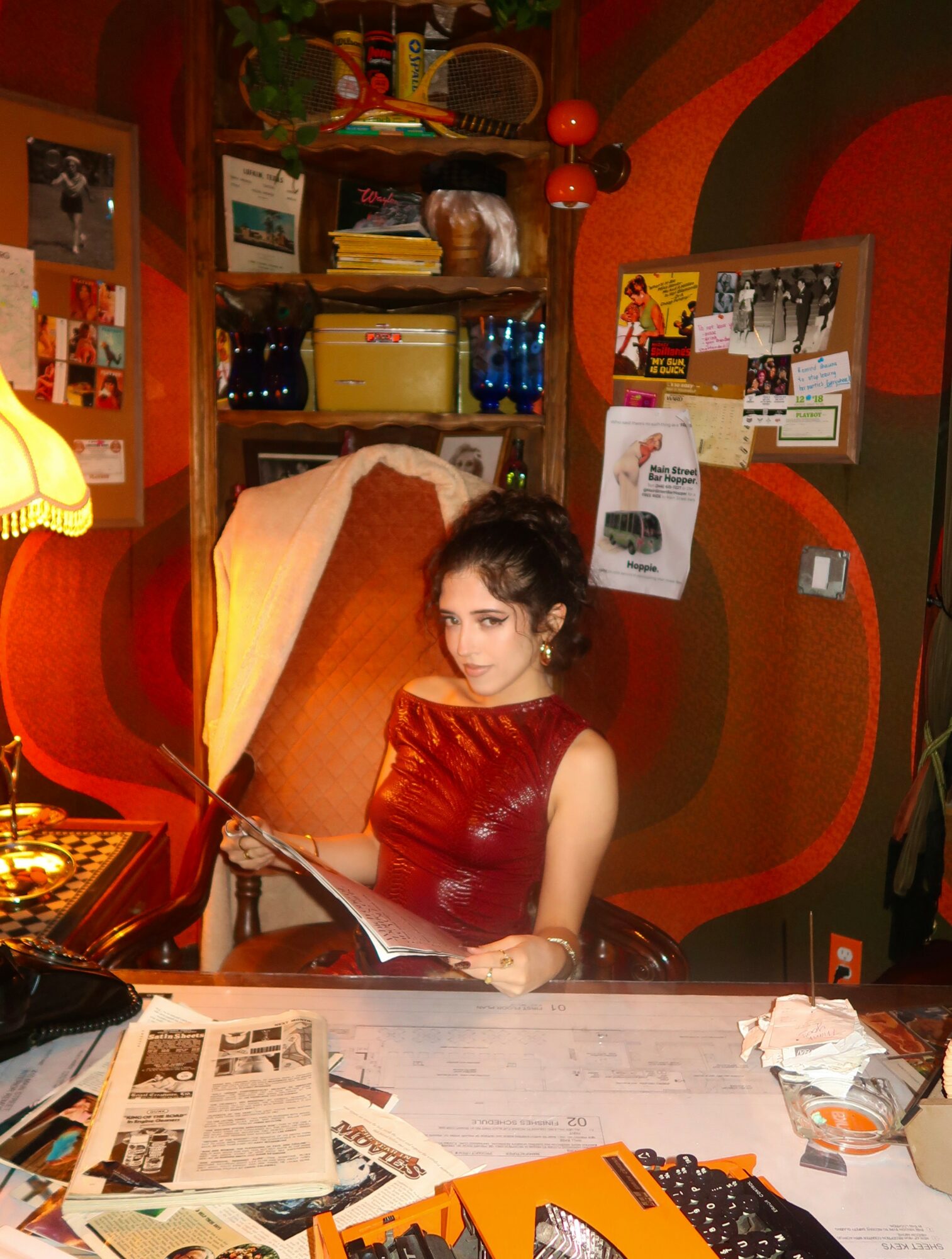 Young woman reading a newspaper at a cluttered desk in a colorful room with shelves and posters.