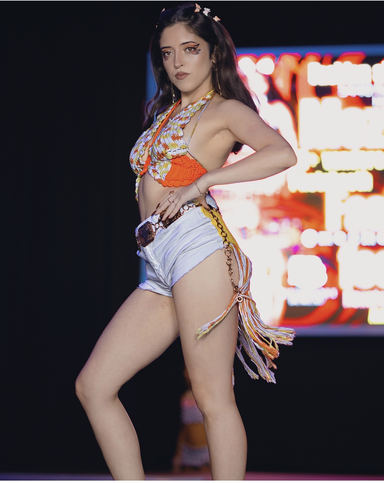 Young woman with long dark hair in colorful top and denim shorts poses on stage with a blurred background.
