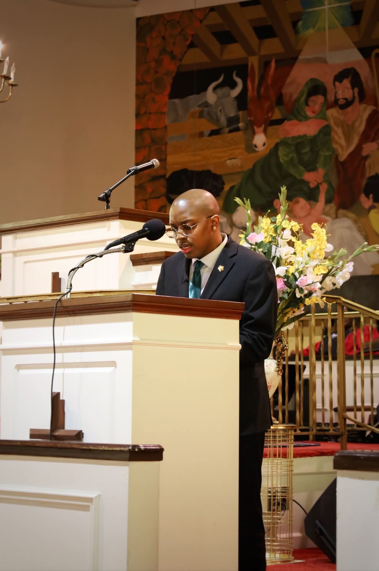 Man in suit speaking at a podium with a microphone, floral arrangement, and a colorful mural in the background.