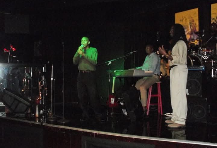 Three people on stage, one singing into a microphone, others playing instruments, dark background, stage lighting.