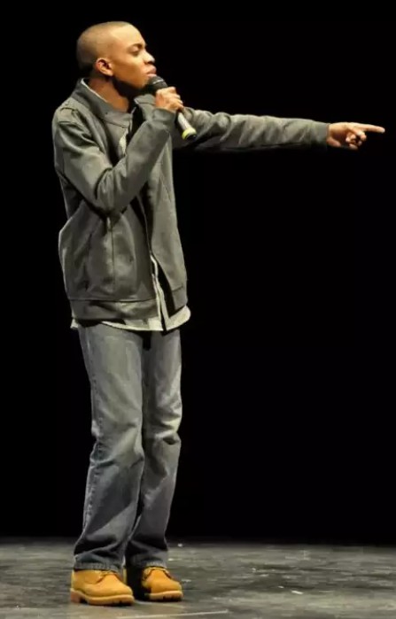 Person standing on stage, holding microphone, pointing to the right, wearing gray jacket and jeans, dark background.