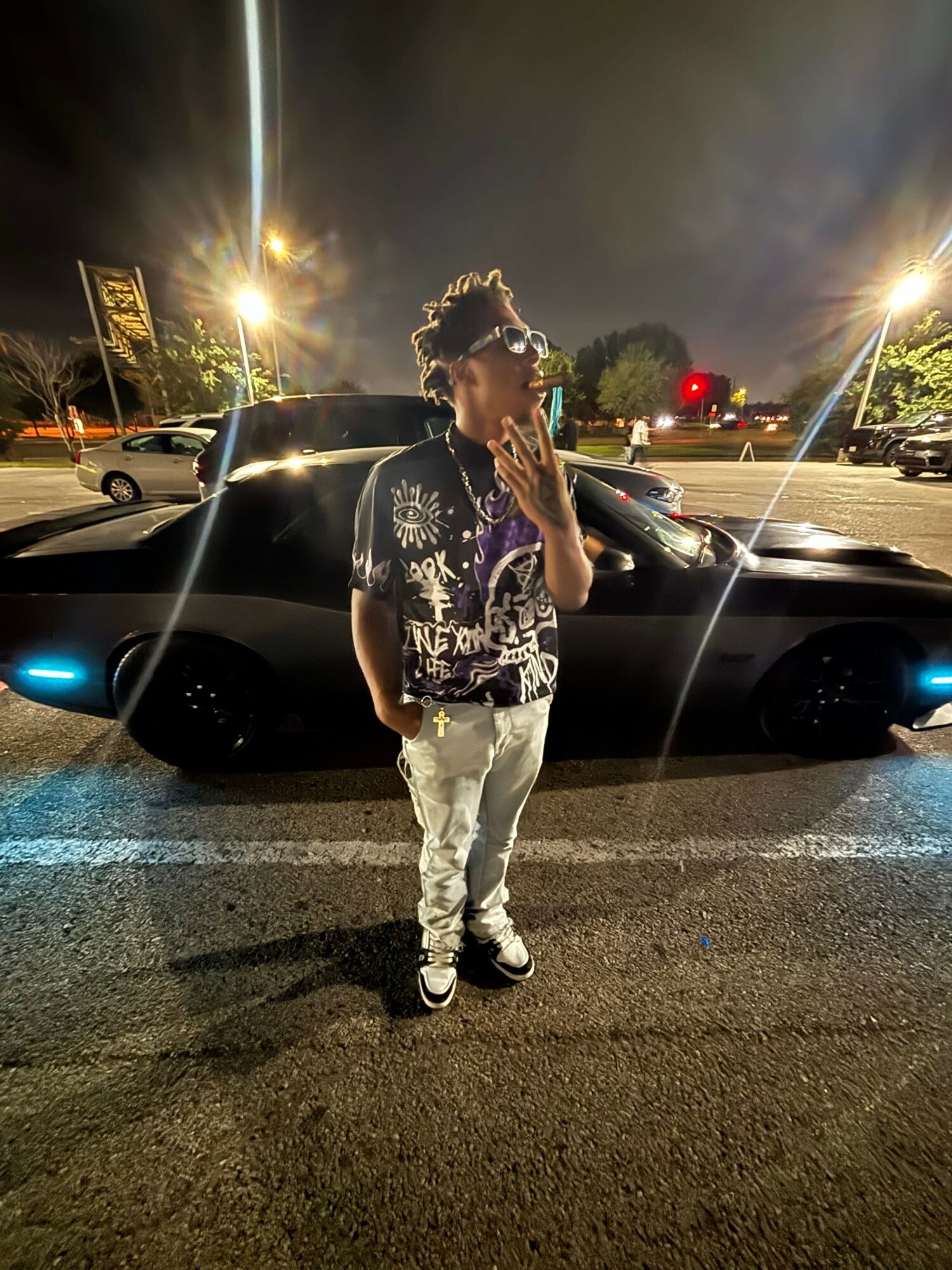 Person standing on street at night, wearing patterned shirt and white pants, with cars and streetlights in background.