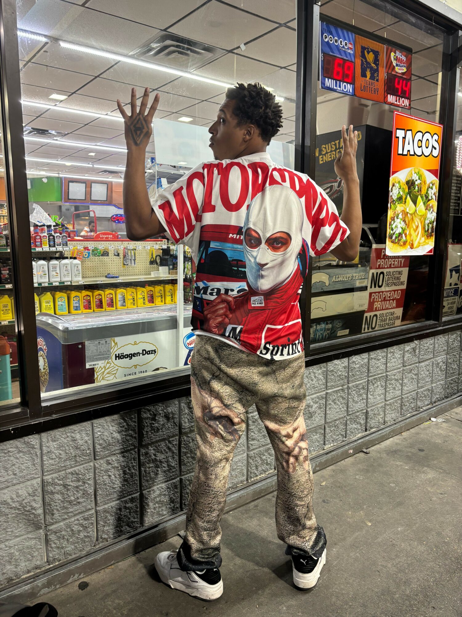 Person standing outside a store window, waving, wearing a red and white shirt, camouflage pants, and sneakers.