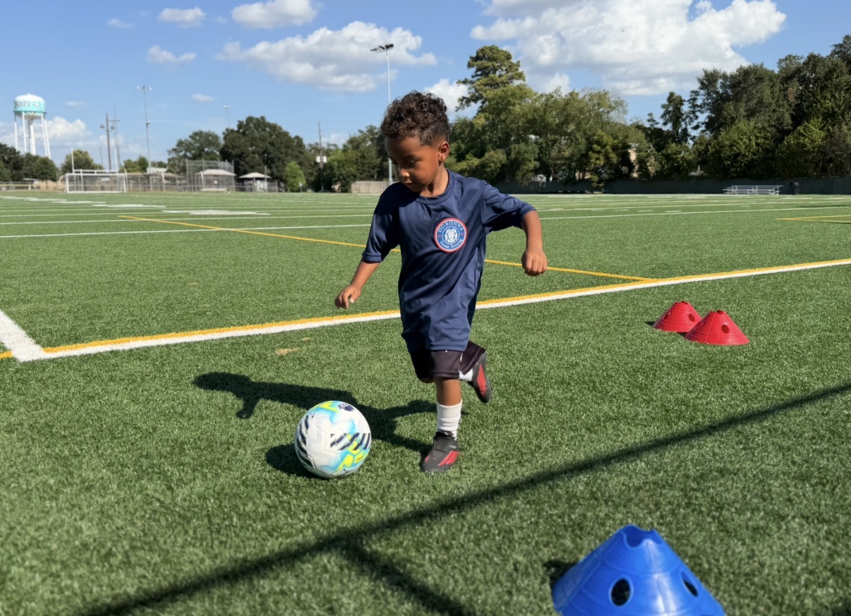 Child playing soccer on a field with cones, kicking a ball under a blue sky with clouds.