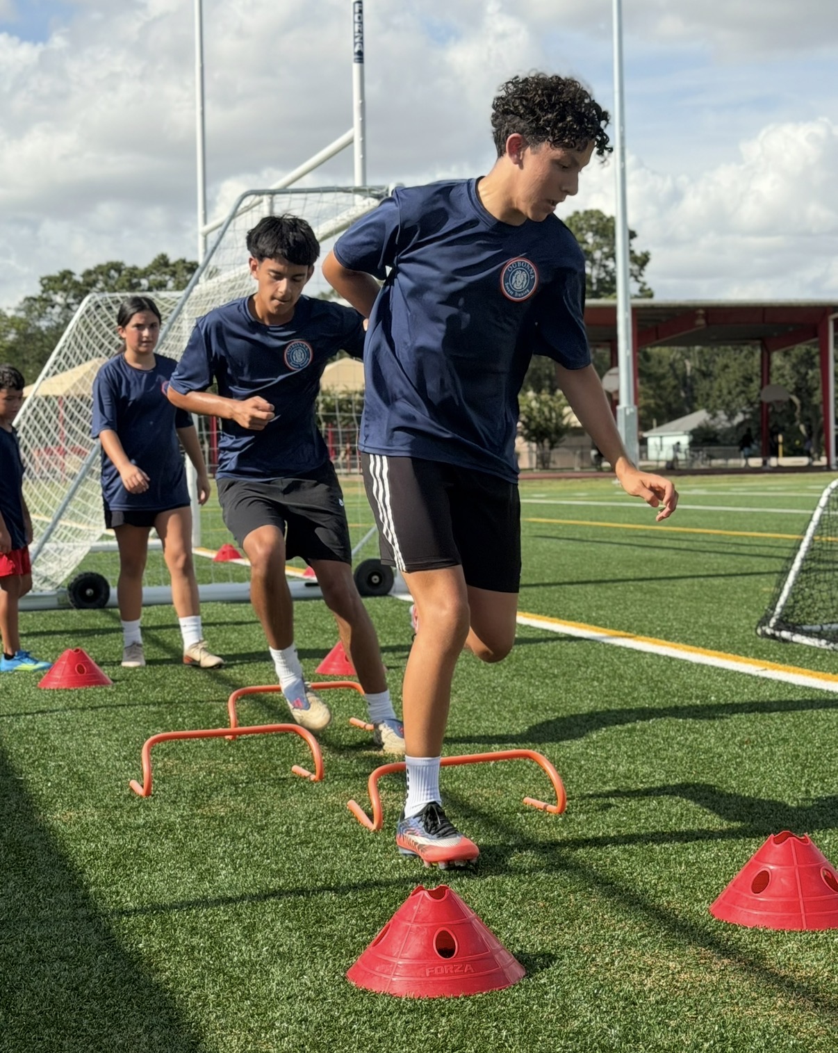 Group of young boys participating in an outdoor athletic training drill on a grassy field, with cones and hurdles.