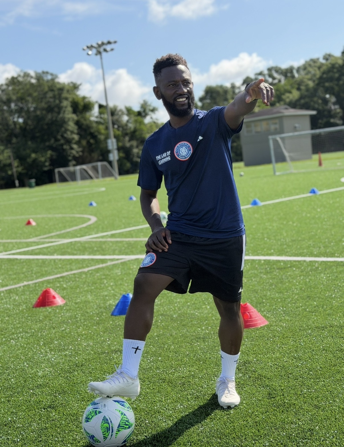 Person standing on a soccer field, pointing with one arm, surrounded by training cones, with a soccer ball at their feet.