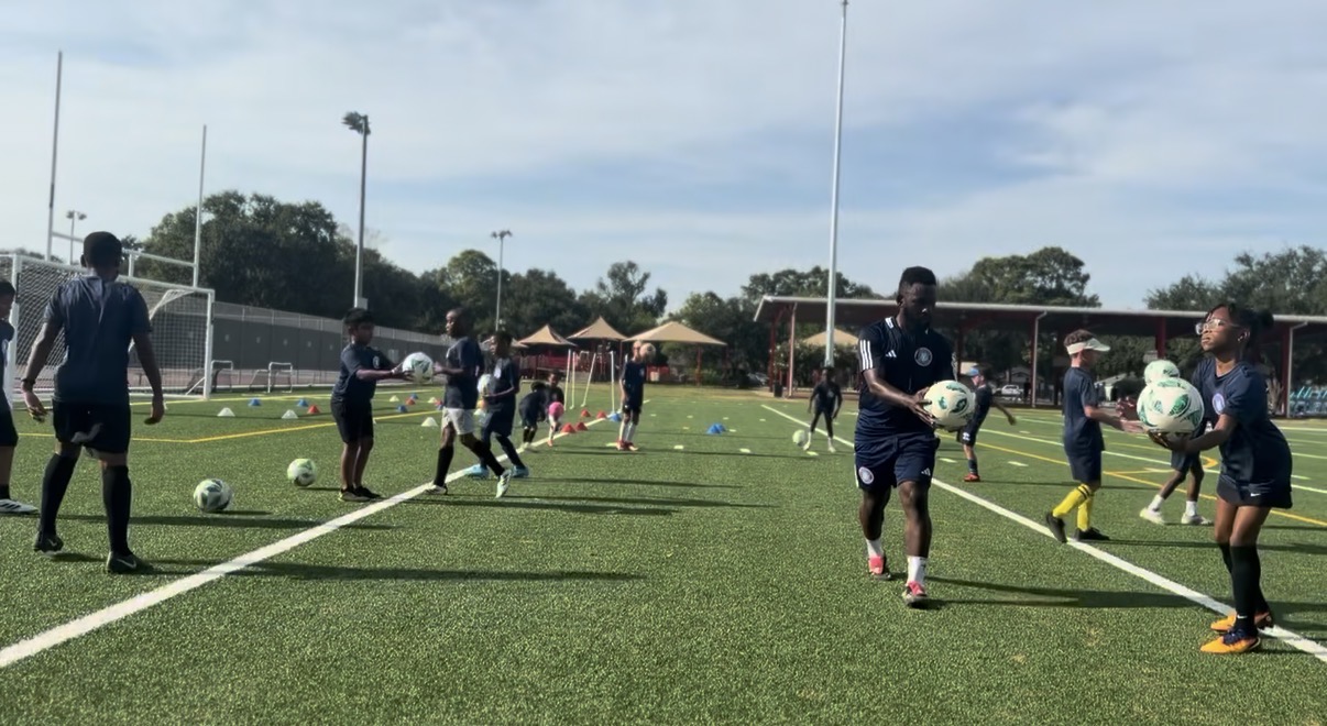 Group of people on a soccer field practicing with soccer balls, some running and others standing, under a partly cloudy sky.