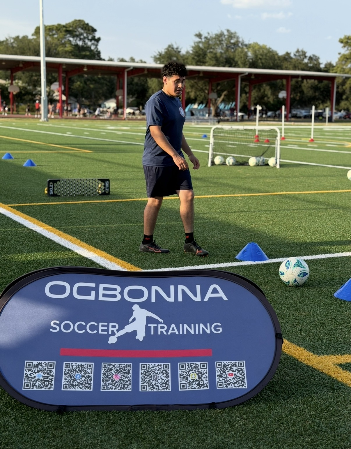 Young person standing on a soccer field with training equipment, soccer balls, and a training sign, outdoors on a sunny day.