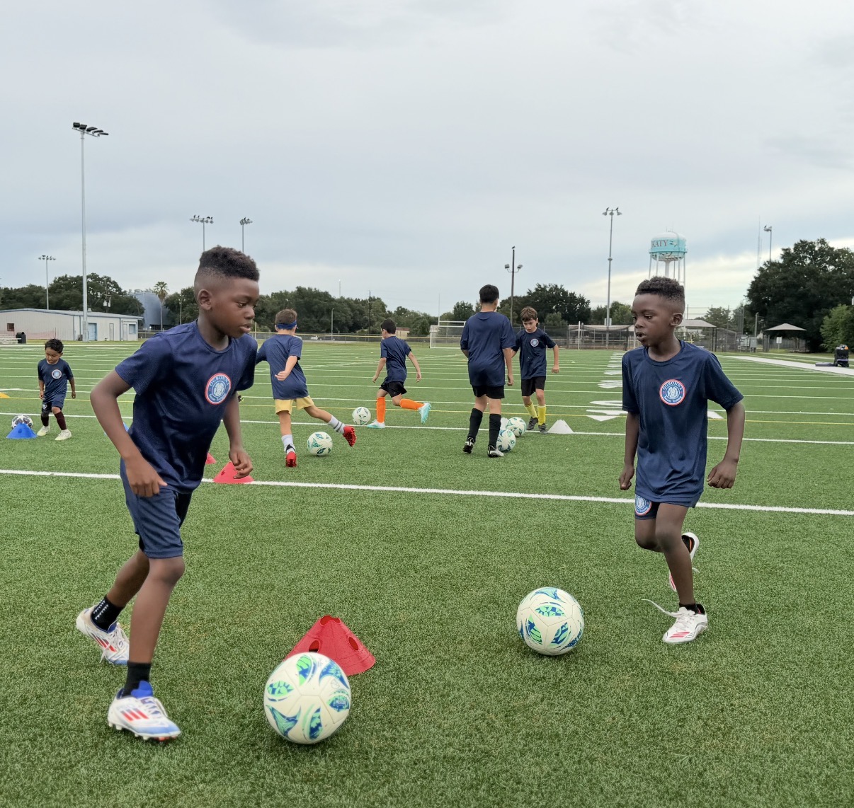 Children in sports uniforms practicing soccer on a field with balls and cones, overcast sky, trees, and light poles in background.