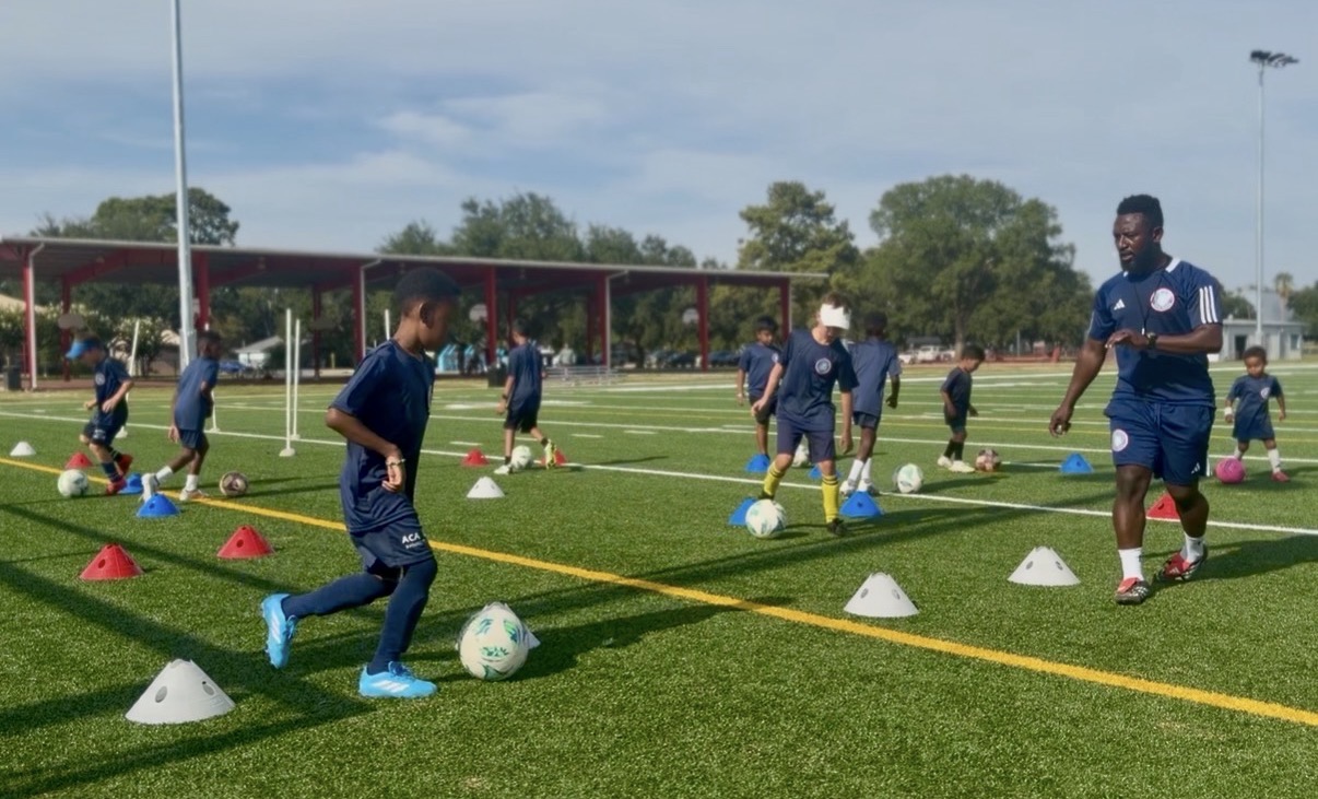 Group of children and coach practicing soccer drills on a field with cones and balls, under a blue sky.