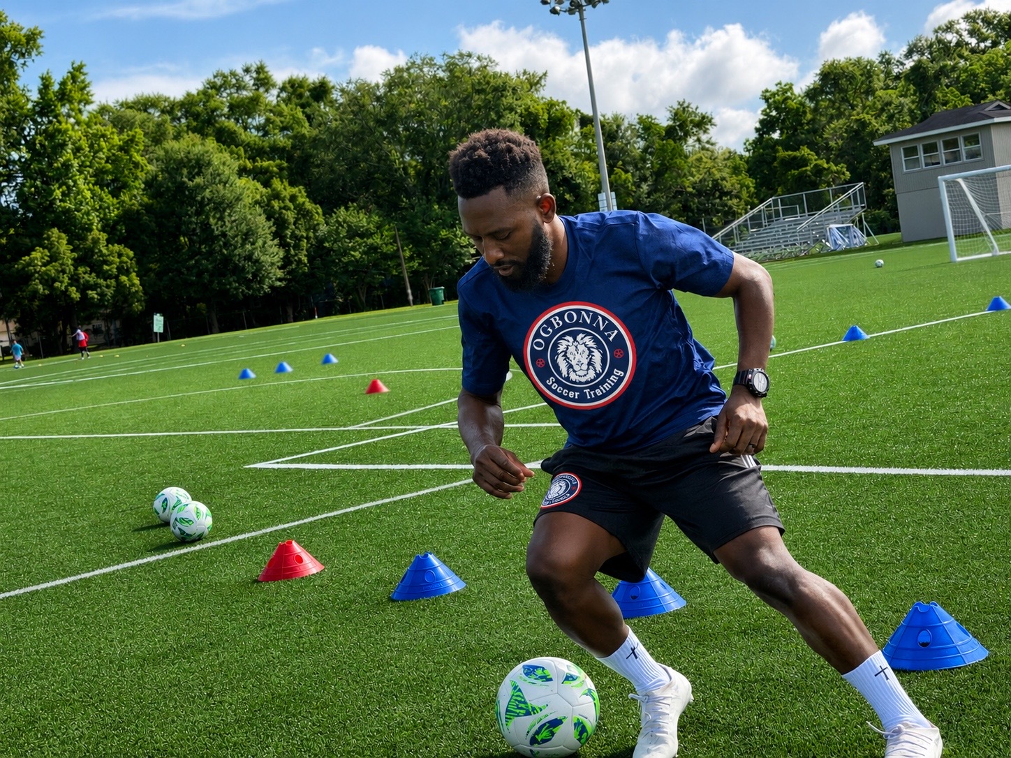 Young man in a blue shirt and black shorts dribbling a soccer ball on a green field with training cones, trees, and a building in background.