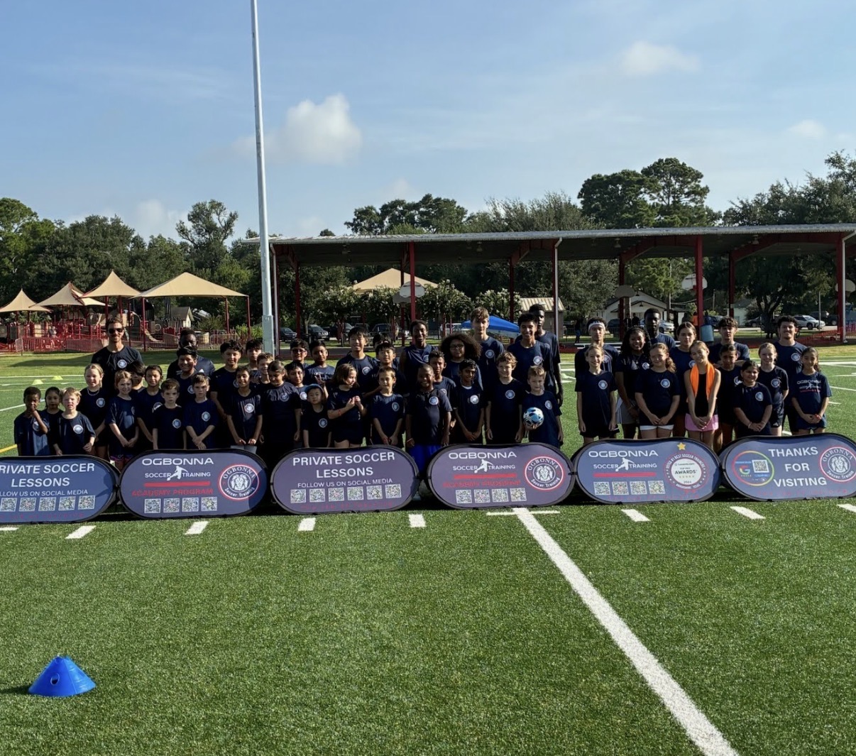 Group of children and coaches on a soccer field holding banners, with trees and tents in the background.