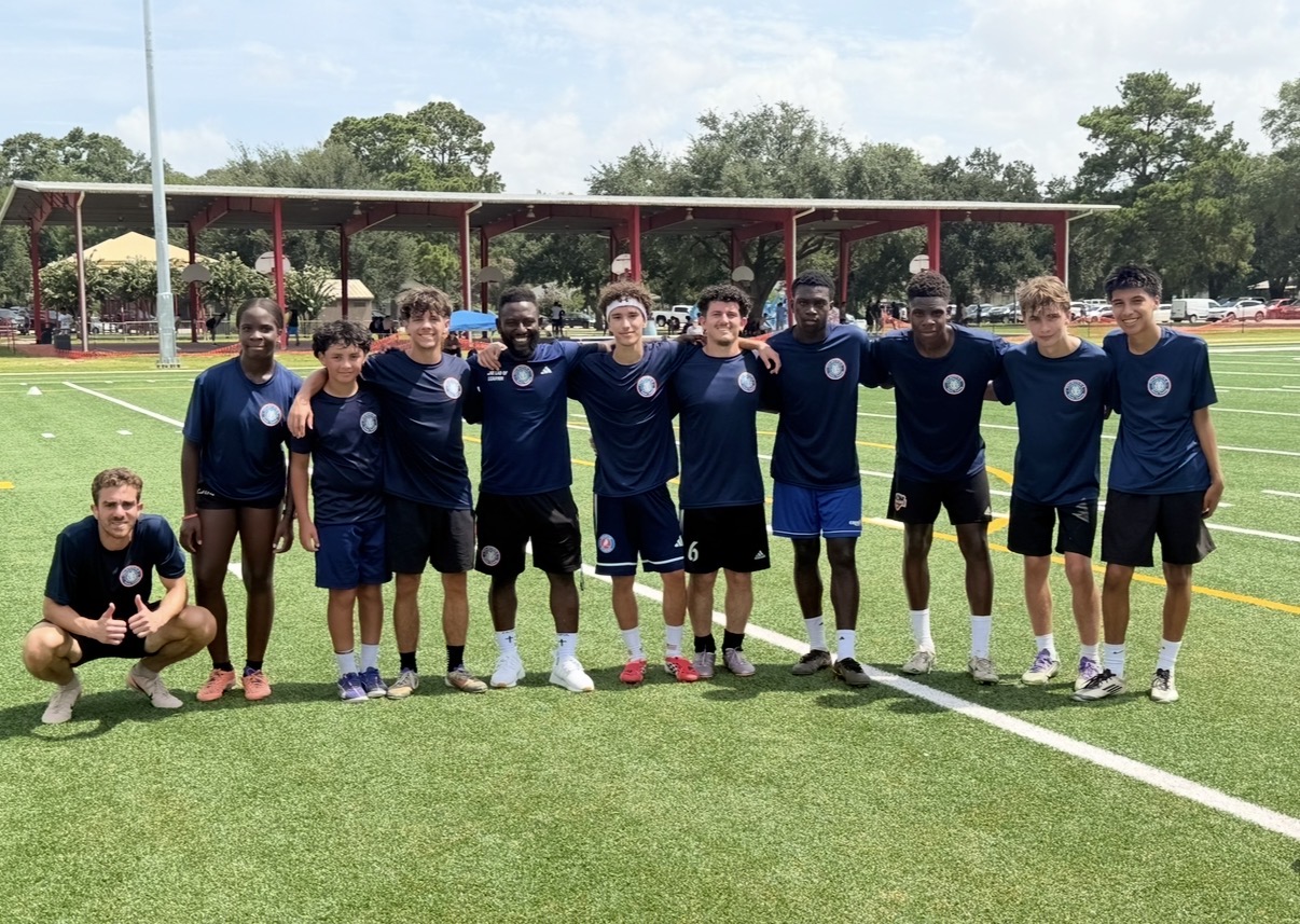 Group of nine young people standing on a sports field, some with arms around each other, wearing matching dark blue sports uniforms.