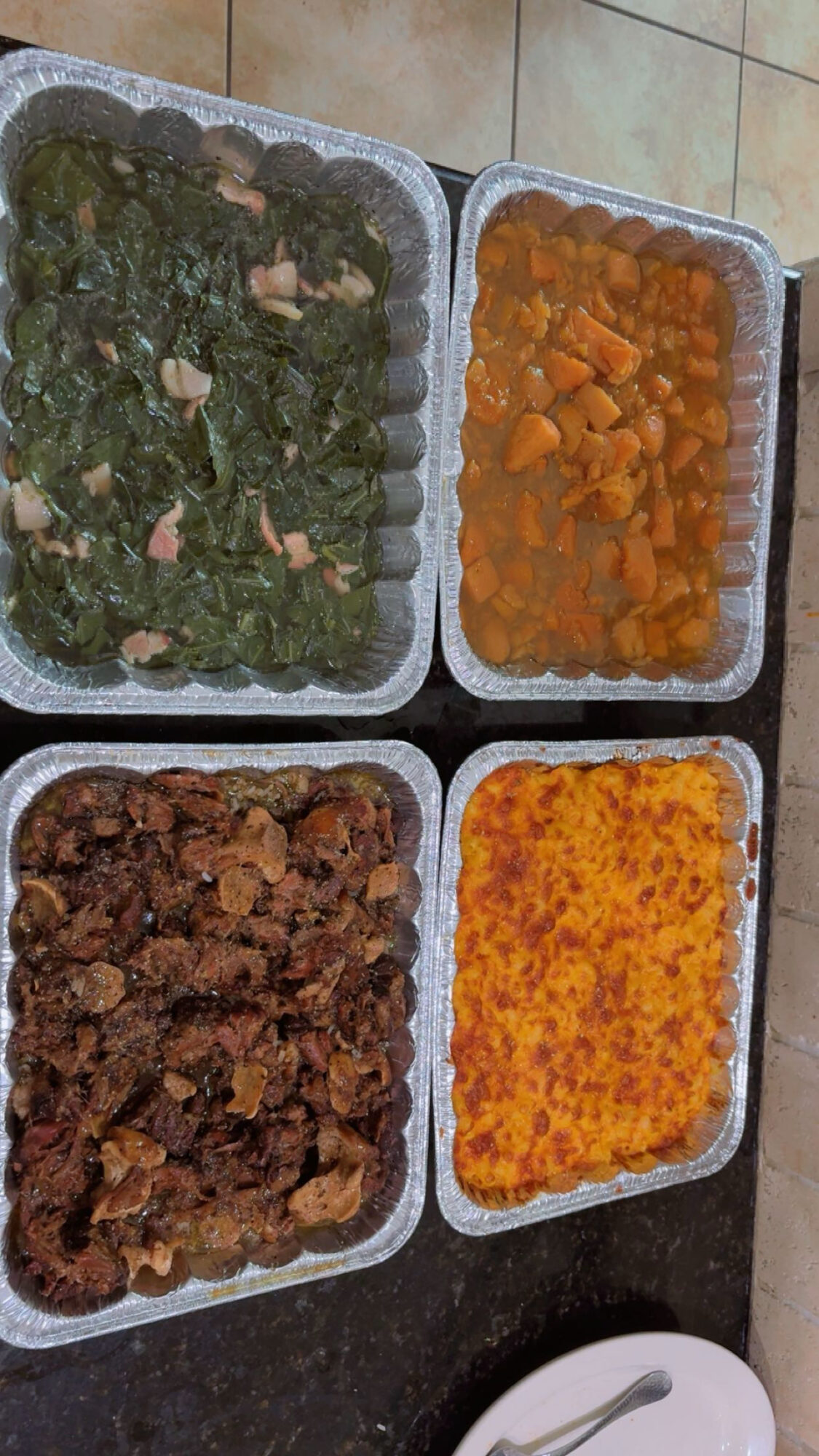 Four trays of cooked food on a black surface, with a white spoon nearby. The trays contain green, orange, brown, and yellow dishes.