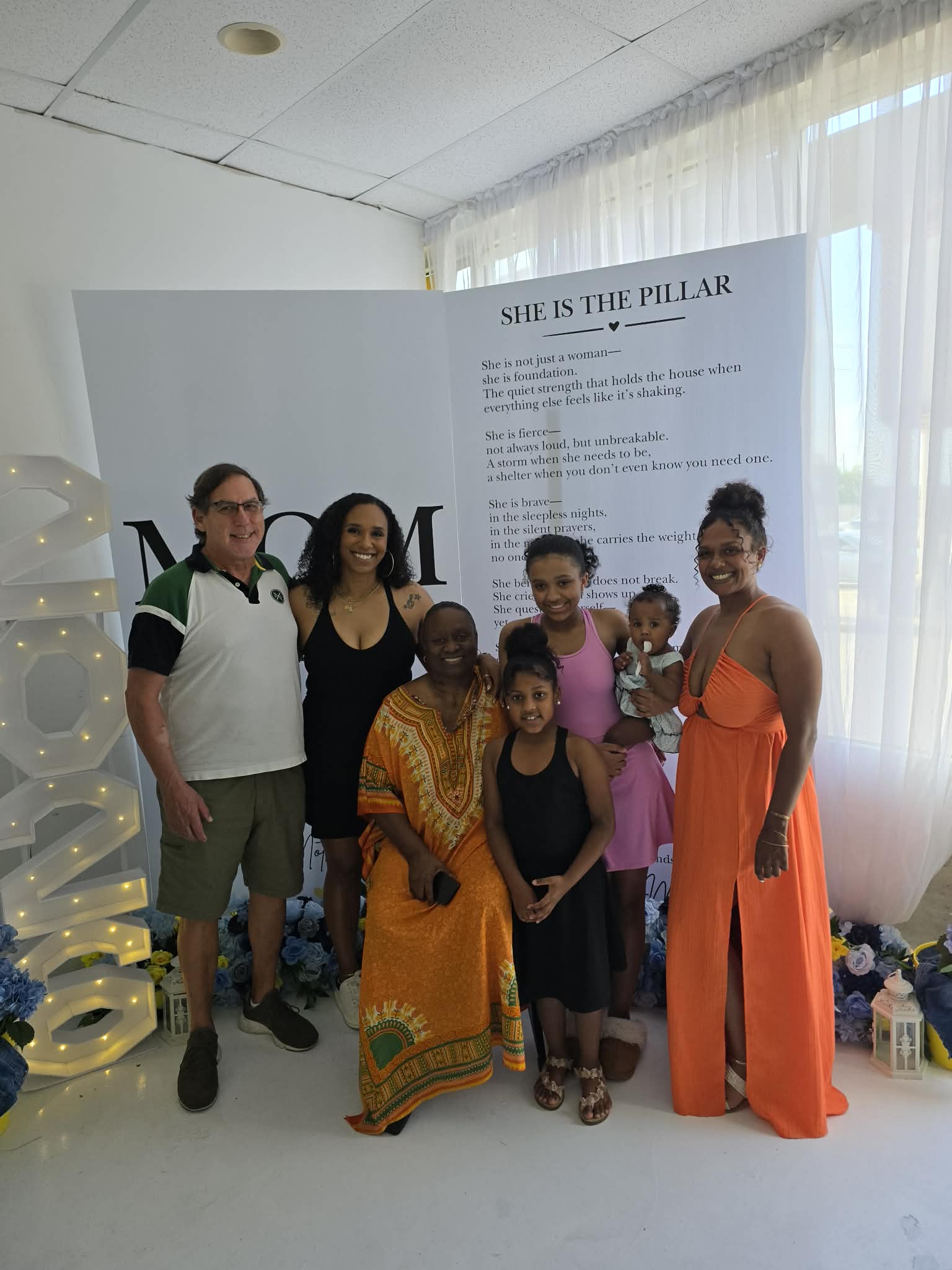 Group of seven people, including children, standing indoors near a white wall with text, smiling for a photo.