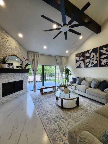 Living room with a ceiling fan, large window, sofa, artwork, and a fireplace, decorated with plants and a rug.