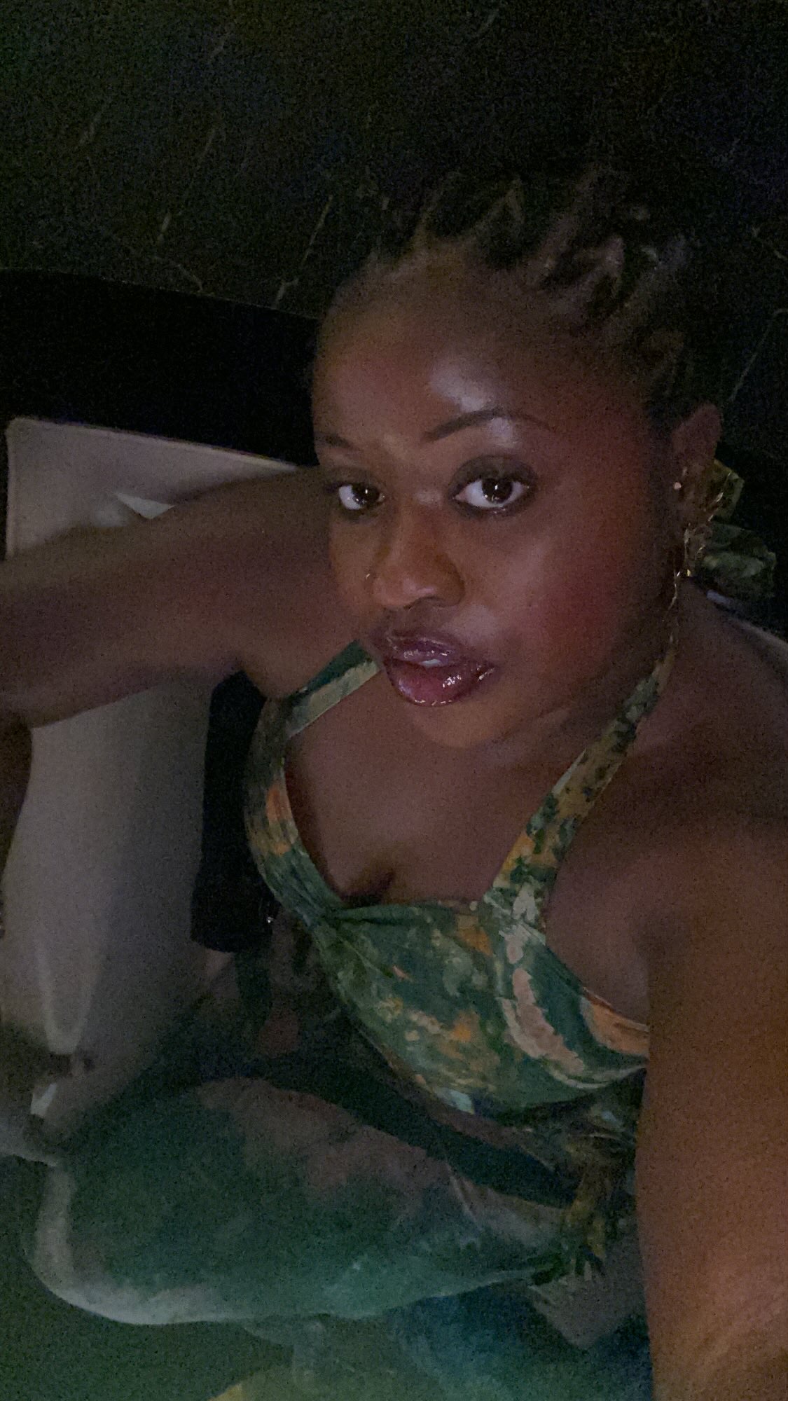 Woman with braided hair wearing a patterned top, looking at the camera, in a dimly lit setting.