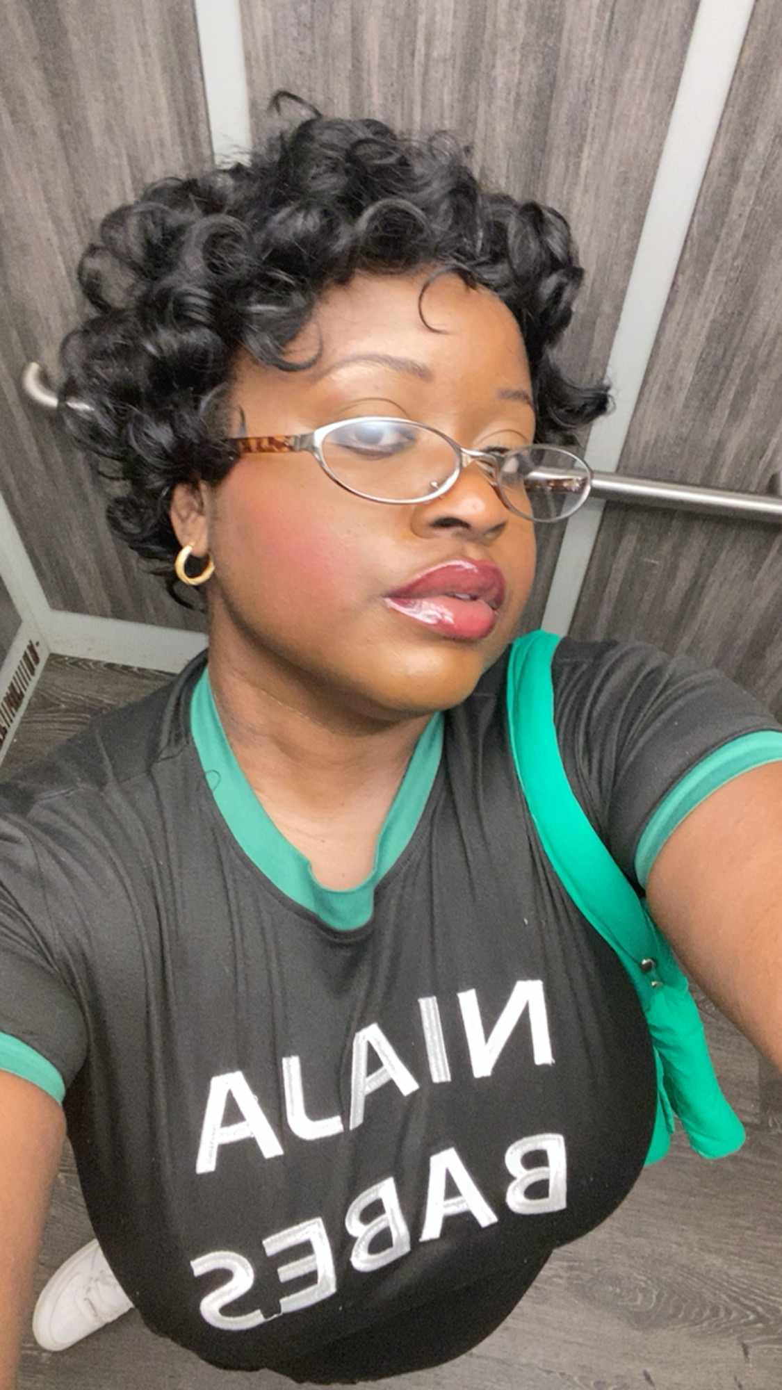 Woman with short curly hair wearing glasses and a black T-shirt with green accents, carrying a green bag, in an indoor setting.