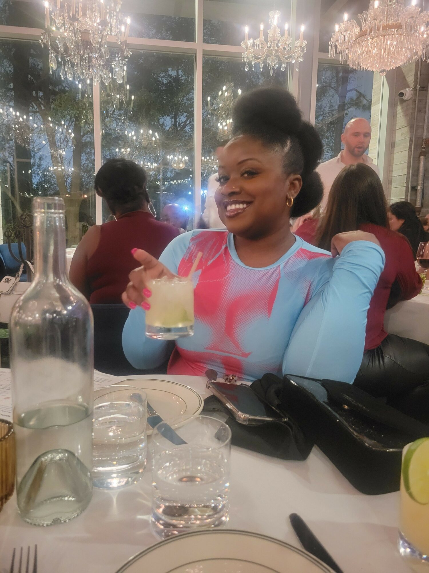 Person smiling and holding a slice of cake at a dining table with glasses and a bottle, in a decorated room.