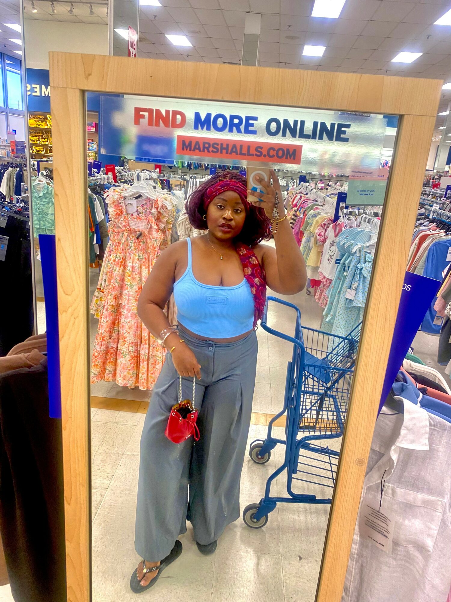 Woman taking a mirror selfie in a store, wearing a blue top, gray pants, and a red headscarf, with shopping carts behind her.