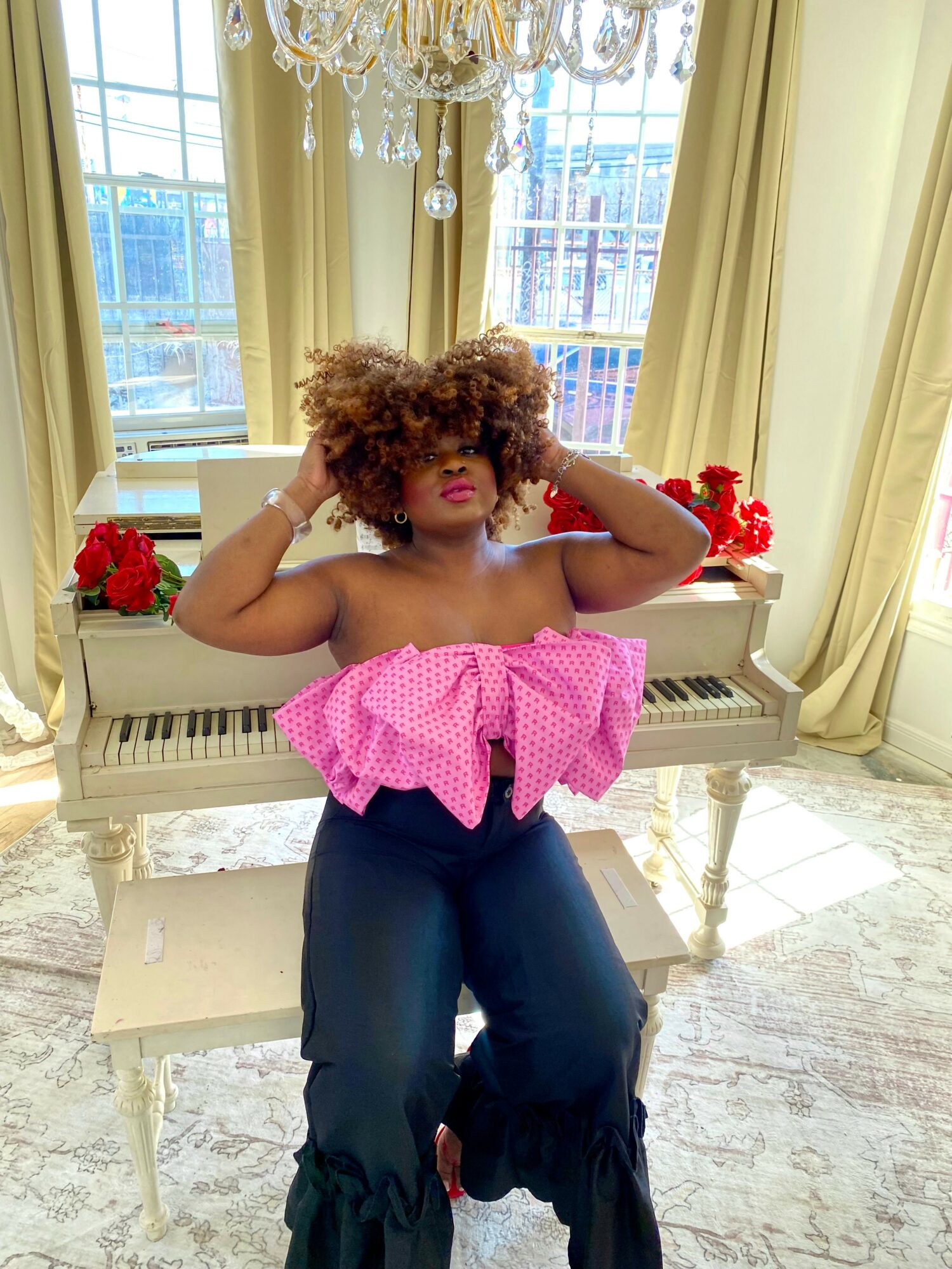 Child with curly hair in pink top and dark pants standing indoors near windows with curtains and a chandelier.
