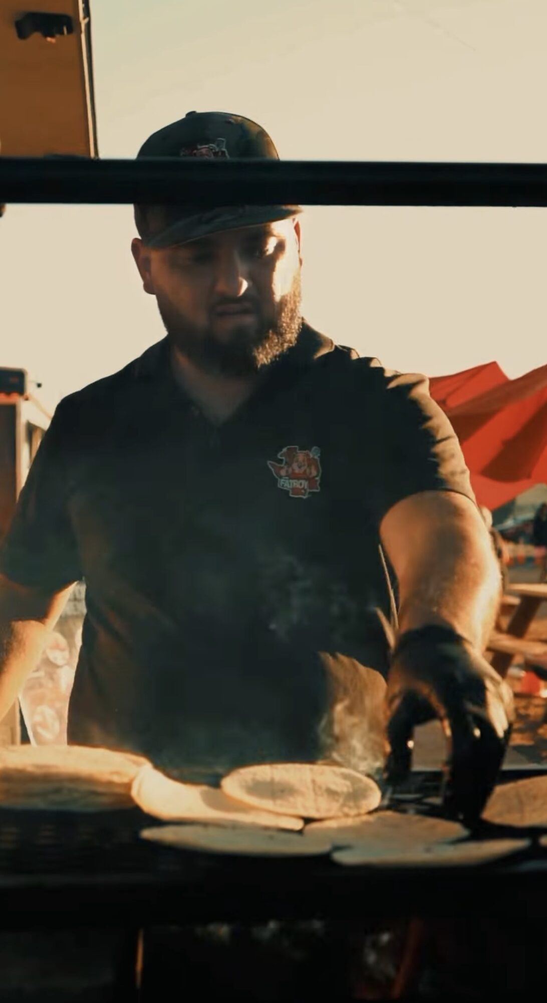 Man with beard wearing a cap and black shirt, cooking outdoors with smoke rising, red umbrella in background.