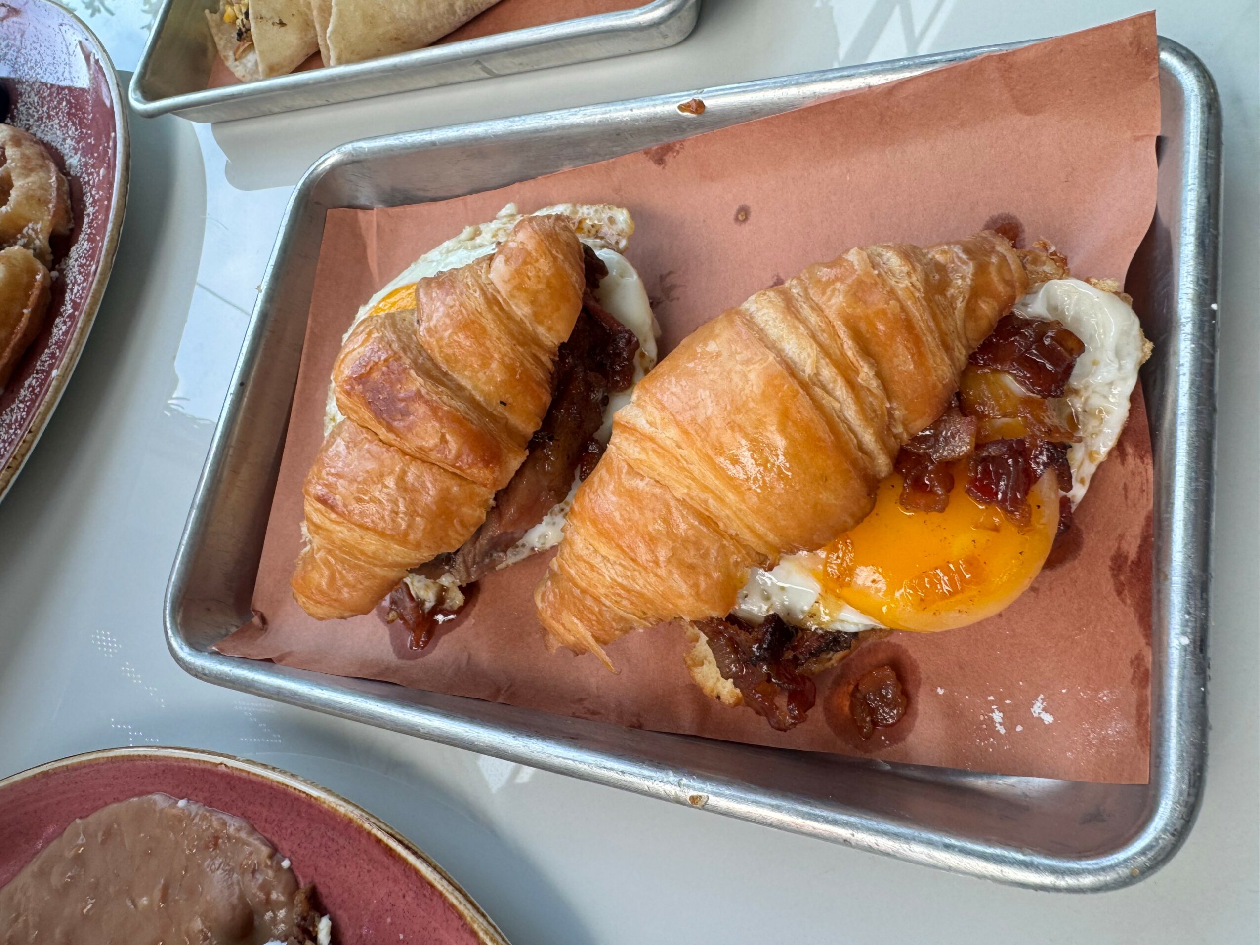 Two croissant sandwiches with eggs, bacon, and other fillings on a metal tray with pink paper lining.