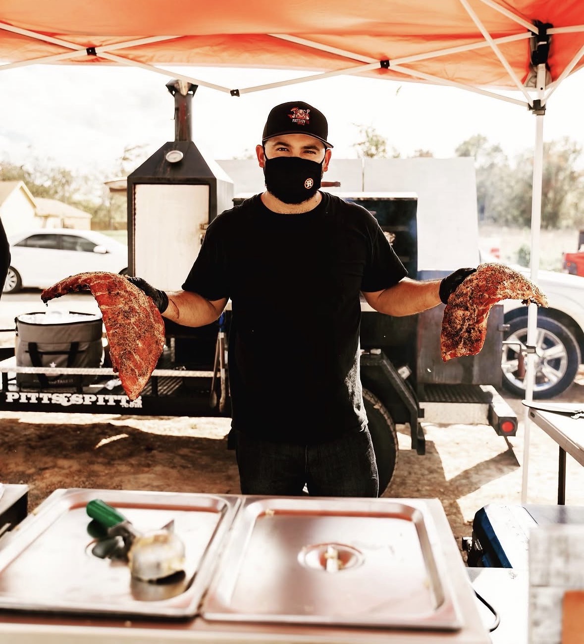Person wearing a black mask and black shirt holding two large pieces of cooked meat outdoors under an orange canopy.