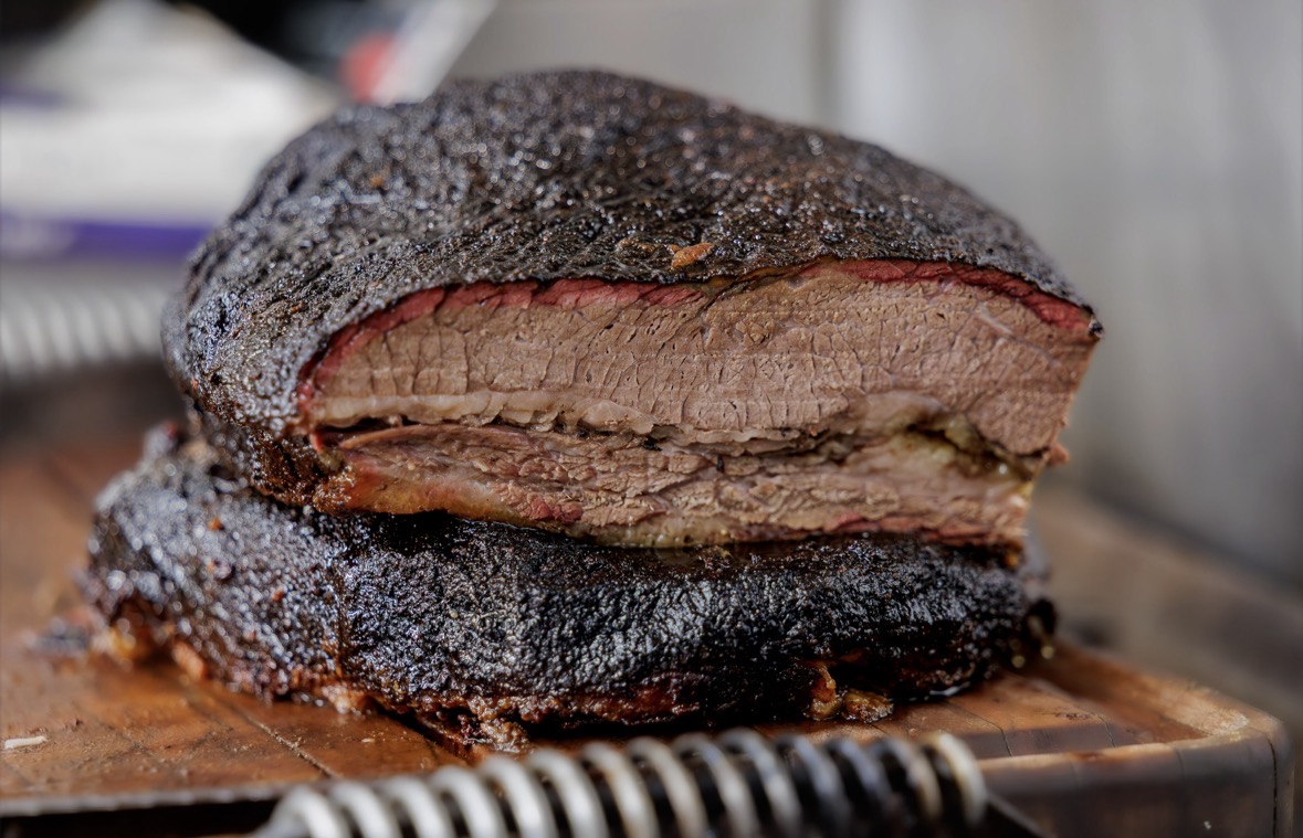 Cooked beef brisket with a dark crust, sliced to show tender meat inside, on a wooden surface.